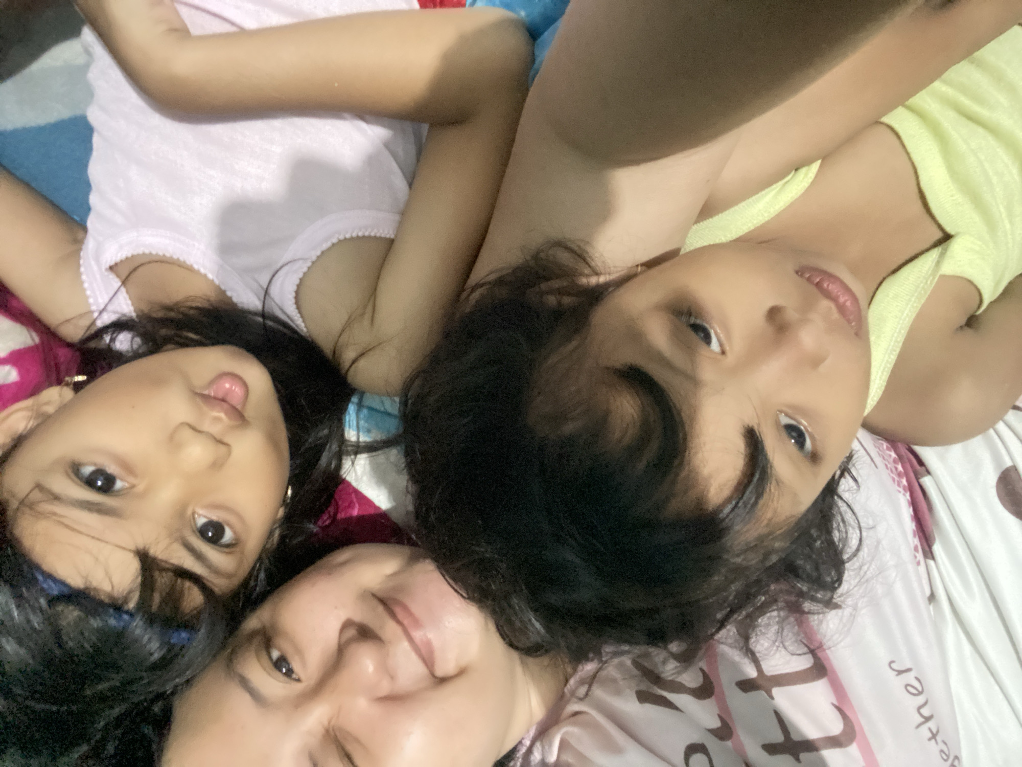 This is a close-up, overhead shot of three individuals, likely a mother and two children, lying down together on a bed. The two children are positioned at the top of the frame, with their faces tilted towards the camera. The youngest child, on the left, has dark hair and is looking directly at the camera with her tongue sticking out slightly. The other child, on the right, also has dark hair and is looking sideways with a faint smile. The mother is at the bottom of the frame, with her eyes closed and a smile on her face.
The subjects are dressed casually, with the children wearing what appear to be tank tops. One child is wearing a light pink top, and the other a light yellow top. The mother is wearing a pink top. They are lying on a bed with a patterned sheet. The sheet has a repeating pattern of circular shapes and the word "together" in script. The time of day appears to be evening or night, as the lighting is warm and subdued. The setting seems to be indoors, in a bedroom. The overall mood of the image is one of warmth, affection, and togetherness. There are no external environmental cues visible.