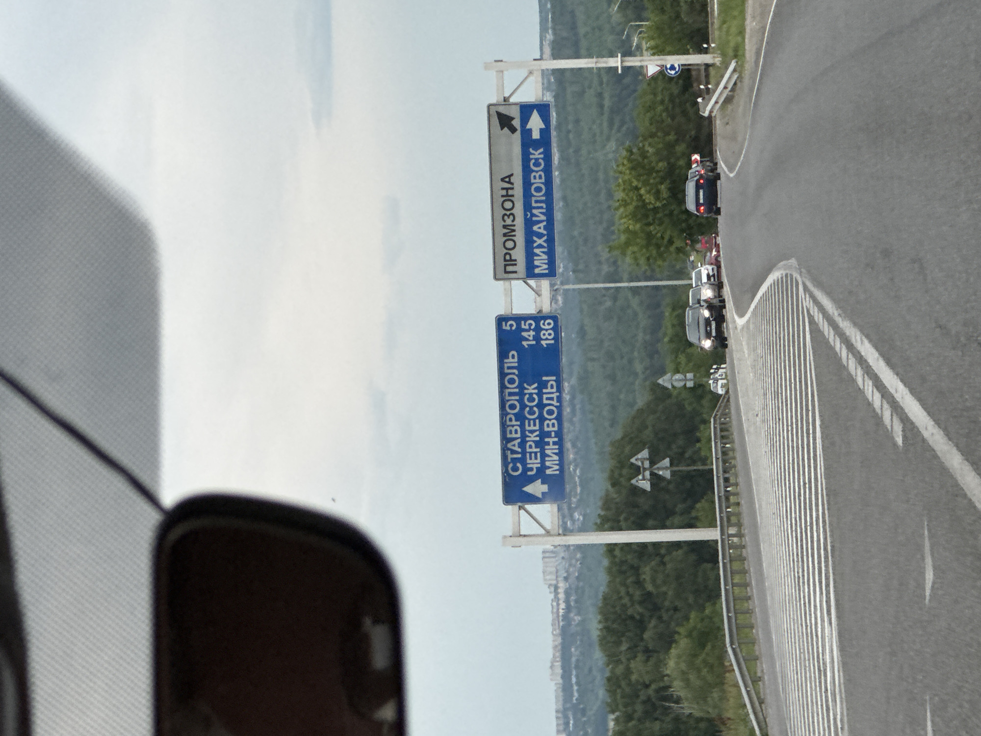 This media file is a photograph taken from inside a vehicle, likely a car, looking out at a highway scene. The image is oriented vertically. The primary subjects are road signs, a highway, and a line of cars.
There are two prominent road signs. The upper sign is rectangular and has two sections. The left section is a light grey with the Russian text "ПРОМЗОНА" (Promzona) in black, indicating an industrial zone. An arrow pointing left is also present. The right section of this sign is blue with white Russian text "МИХАЙЛОВСК" (Mikhaylovsk) and a white arrow pointing right.
Below this is another blue rectangular sign with white Russian text. The top line reads "СТАВРОПОЛЬ" (Stavropol) with a left-pointing arrow. Below that is "ЧЕРКЕССК" (Cherkessk) with a left-pointing arrow. The bottom line reads "МИН-ВОДЫ" (Min-Vody) with a left-pointing arrow. To the right of these city names are numbers: "5", "145", and "186", likely indicating distances in kilometers.
The scene is a highway with multiple lanes of asphalt and white lane markings. A metal guardrail runs along the side of the road. Several cars are visible on the highway, driving in the same direction as the presumed photographer. Some appear to be SUVs or similar vehicles.
The setting is a rural or suburban area with lush green trees and hills visible in the background. In the far distance, a cluster of buildings suggests a town or city. The sky is overcast with a light grey and white hue, indicating potentially cloudy weather. The time of day appears to be daytime, given the lighting.
The overall impression is a travel scene, with directional information for drivers. The focus is on the road and navigational aids, rather than any specific human emotion or event beyond the act of driving.