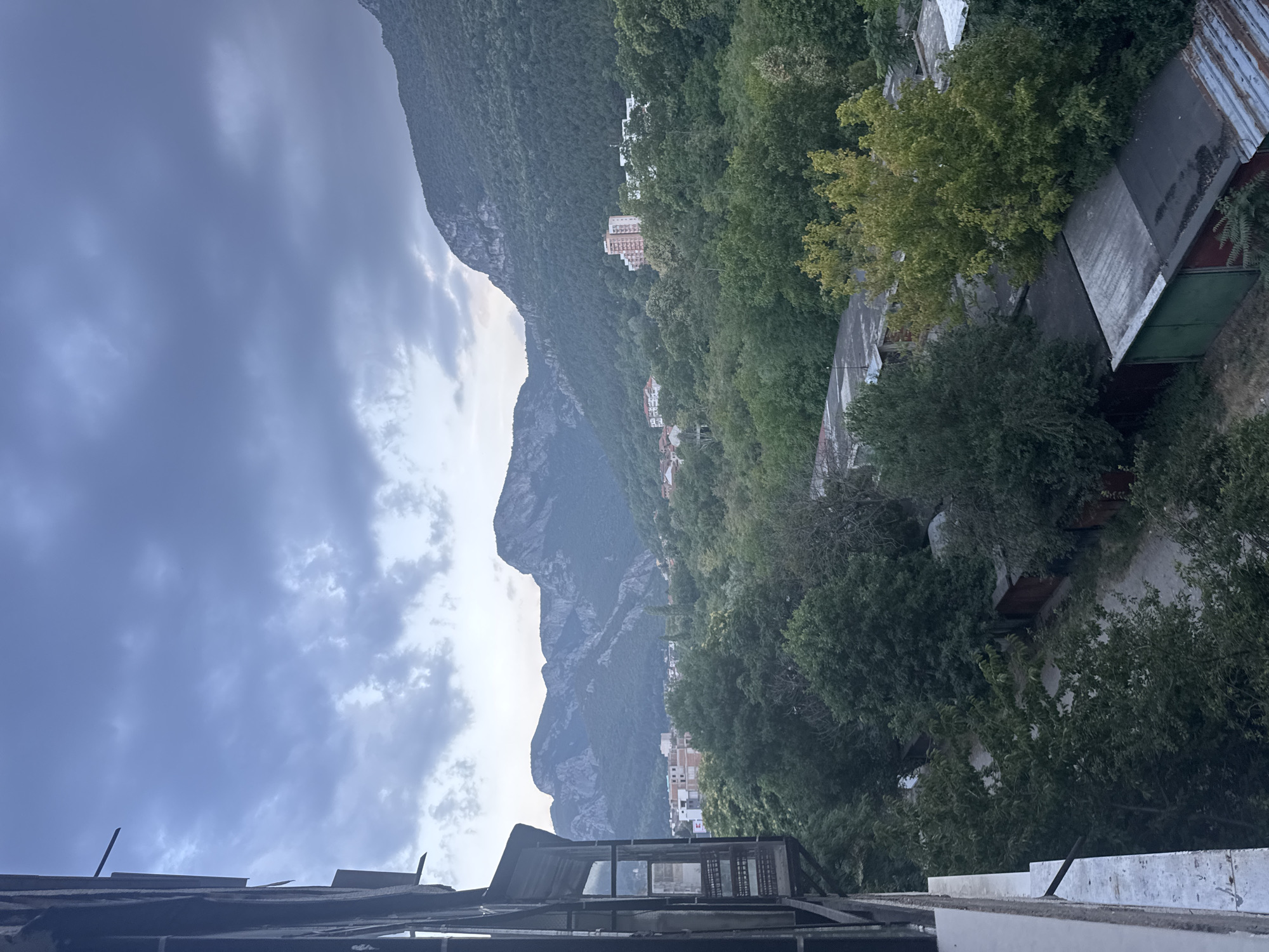 The image shows a view from a balcony overlooking a valley with mountains in the background. In the foreground, there is a portion of a building on the left side, with a balcony. Below the balcony, there are several garages with corrugated metal roofs. A dirt path runs between the garages. Further in the distance, there is a dense forest that covers the lower slopes of the mountains. Scattered within the forest are some buildings, including a few apartment buildings. The mountains are steep and rocky, with some vegetation growing on their slopes. The sky is overcast with clouds, suggesting it might be late in the day or a cloudy day. The overall scene depicts a semi-urban and natural landscape.