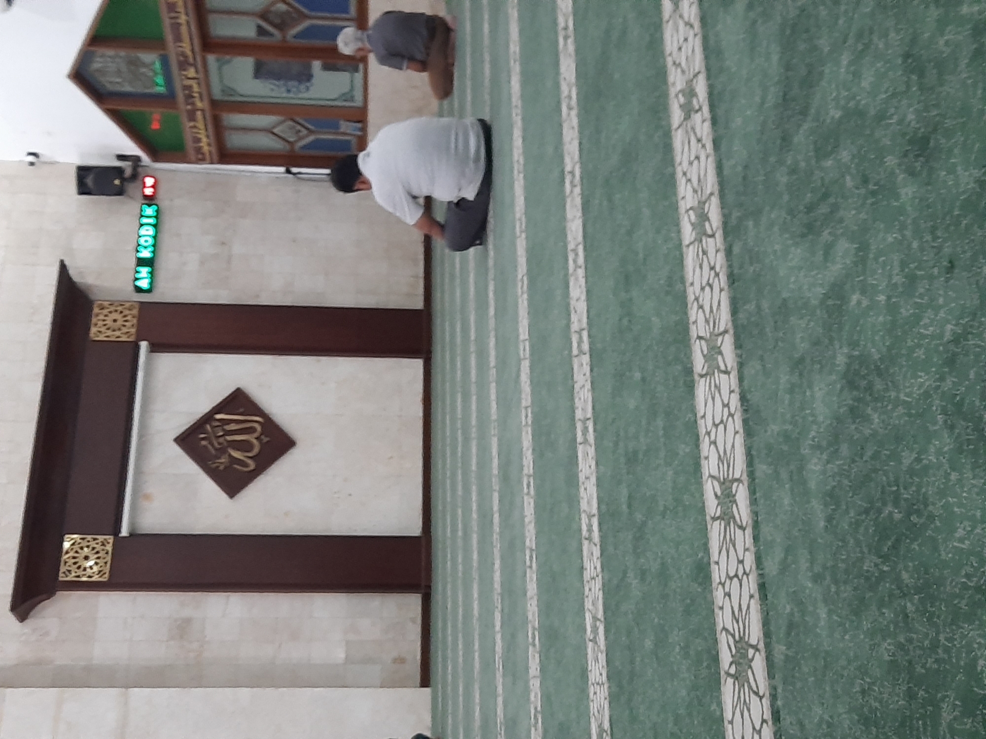 The image shows the interior of a mosque in Bandung City, Indonesia. The floor is covered with a green carpet marked with white stripes. Two men are seated on the carpet, facing away from the camera. One man is wearing a white short-sleeved shirt and dark pants, while the other is wearing a grey shirt and brown pants. In the background, there is a wall with Islamic calligraphy on a diamond-shaped plaque. A digital clock displaying "AH KODIK" is visible on the wall. The mosque has wooden architectural elements and stained-glass windows.