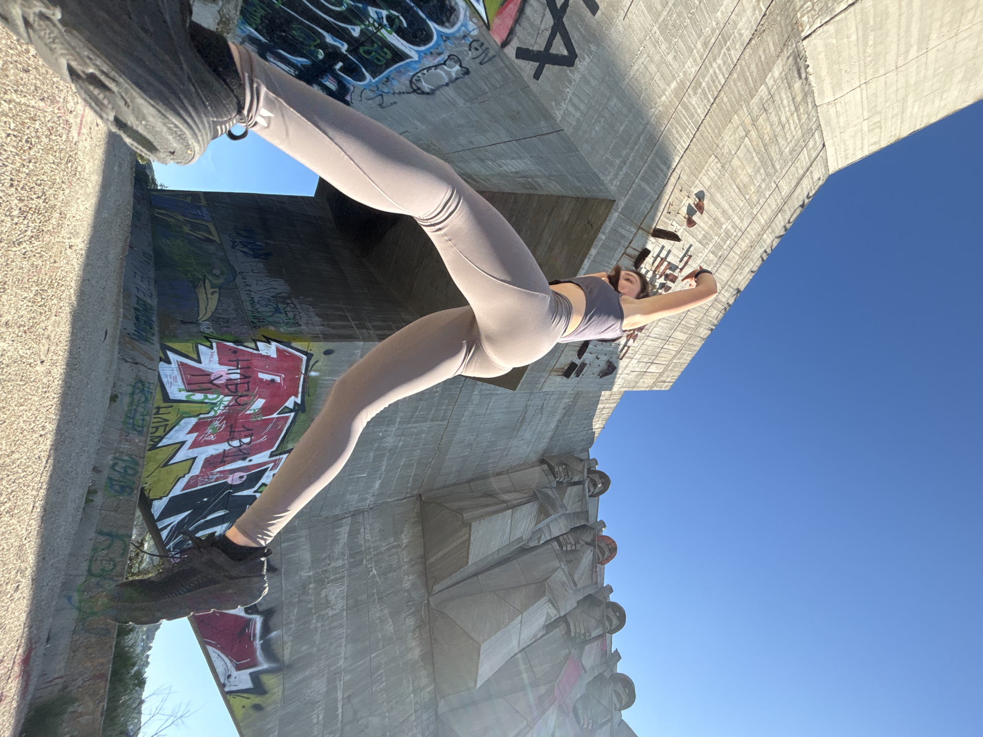 A low-angle shot captures a young woman in athletic wear posing with a vast, Brutalist concrete structure in Varna, Bulgaria. The woman, wearing a grey crop top and light beige leggings, is positioned in the foreground with her right leg extended forward and her left leg bent. Her arms are raised, with her right arm bent at the elbow and her hand reaching upwards, while her left arm is extended outwards. The concrete structure dominates the background, featuring weathered surfaces covered in graffiti. To the right of the woman, at the top of the structure, are several monumental stone figures. The scene is bathed in bright sunlight, casting shadows and highlighting the texture of the concrete and the woman's form. The sky is a clear, vibrant blue. The specific location is the Buzludzha Monument, a now-abandoned former headquarters of the Bulgarian Communist Party, located in the Stara Planina mountains, not in Varna.