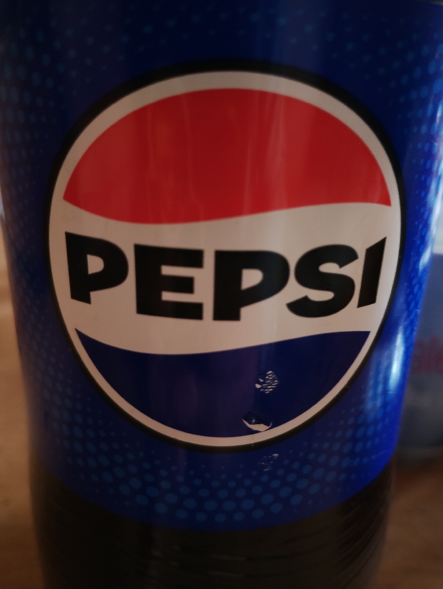 The image shows a close-up of a Pepsi cup. The Pepsi logo, featuring a red, white, and blue circle with the word "PEPSI" in white letters, is prominently displayed. The cup has a blue, patterned background with small, lighter blue circles. There are a few water droplets visible on the lower part of the logo. The cup is sitting on a surface that appears to be a wooden table. The background is blurred, but suggests an indoor environment. There are no people or specific city details visible in the image.