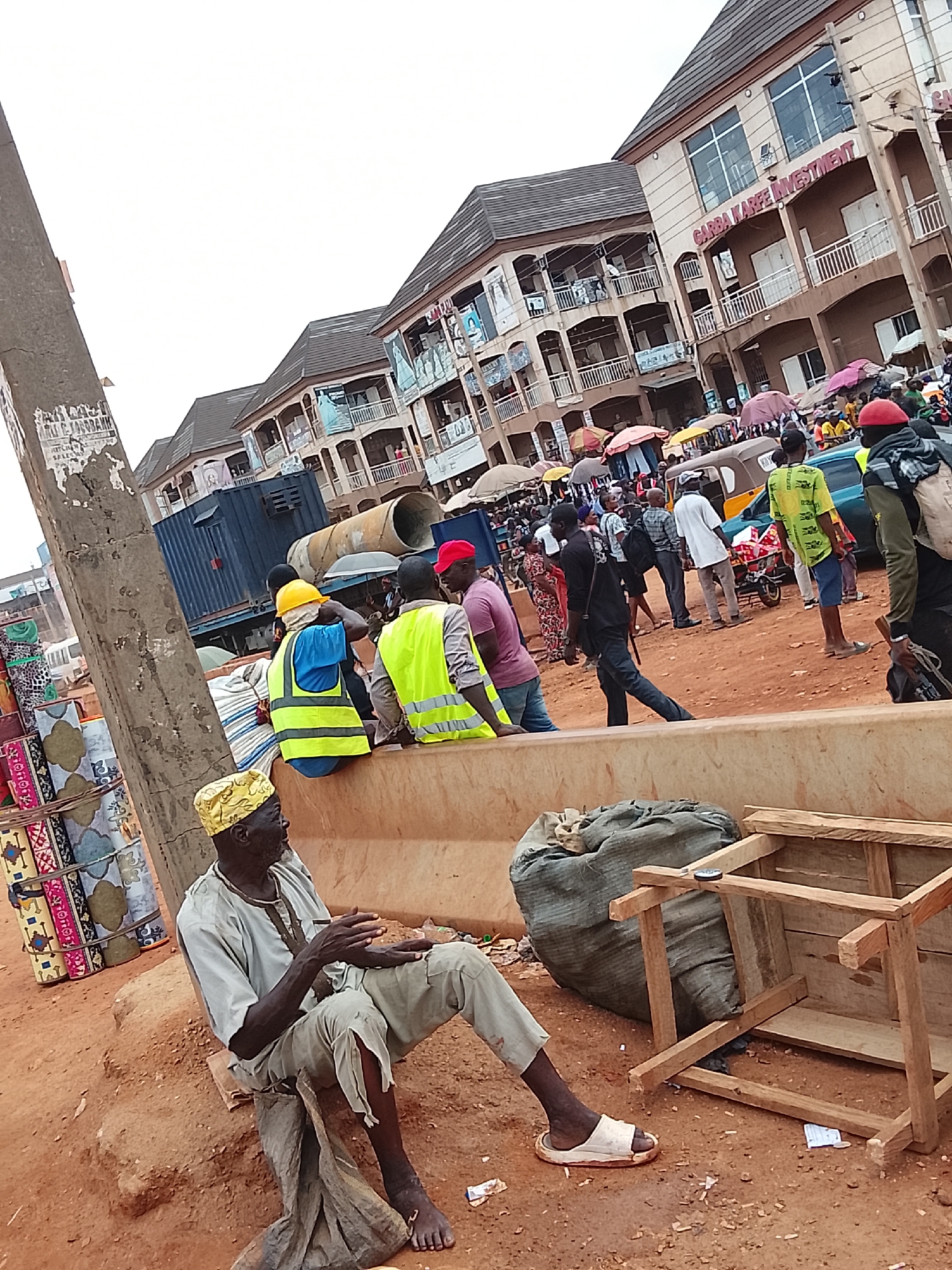 The image depicts a street scene in what appears to be a bustling commercial area. In the foreground, an elderly man wearing a yellow hat, light shirt, and torn pants is seated barefoot on what may be a makeshift seat near a concrete barrier. A wooden frame sits near a large sack next to him. Behind the barrier, several construction workers, dressed in reflective vests and hard hats, are also seated. A crowd of people moves through the area, some carrying umbrellas. In the background, there are several buildings, one of which is labeled "GARBA KARFE INVESTMENT". The ground is dirt or red clay. Based on the request, the location is Katambe, Nigeria.