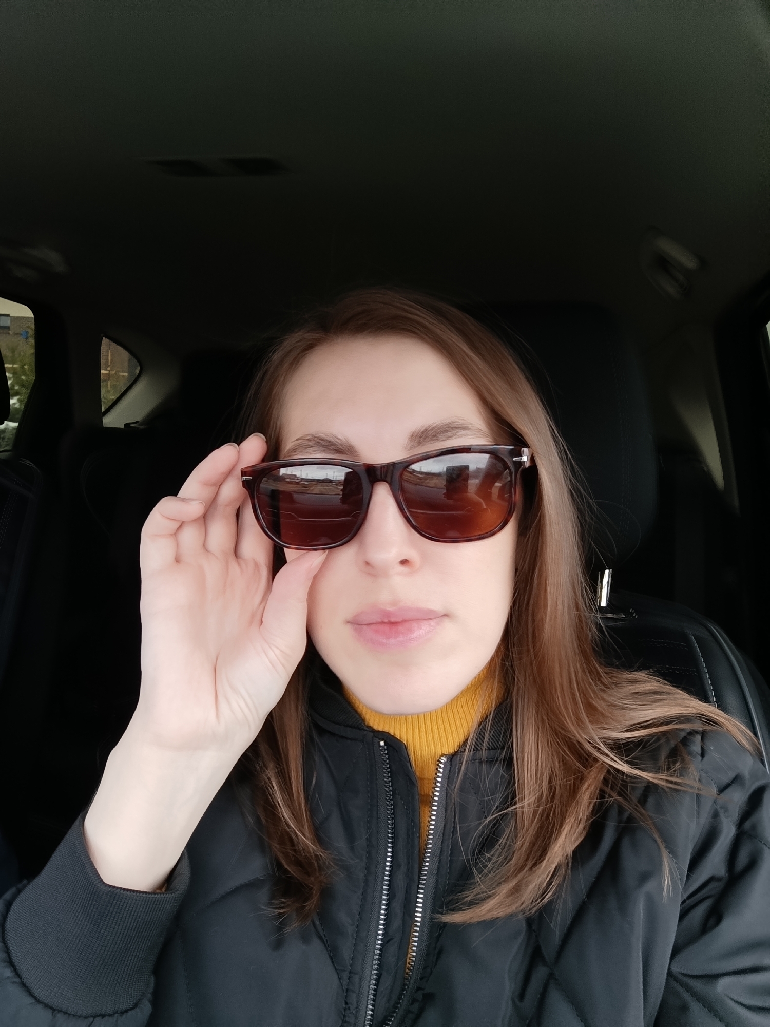 A young woman is in the driver's seat of a car. She is wearing sunglasses, a yellow turtleneck, and a black quilted jacket. She is holding the sunglasses with her left hand. The car's interior is visible, and some buildings are visible through the windshield. The reflection in the sunglasses shows buildings and a cloudy sky. This image was taken in Chaikovsky, Russia.