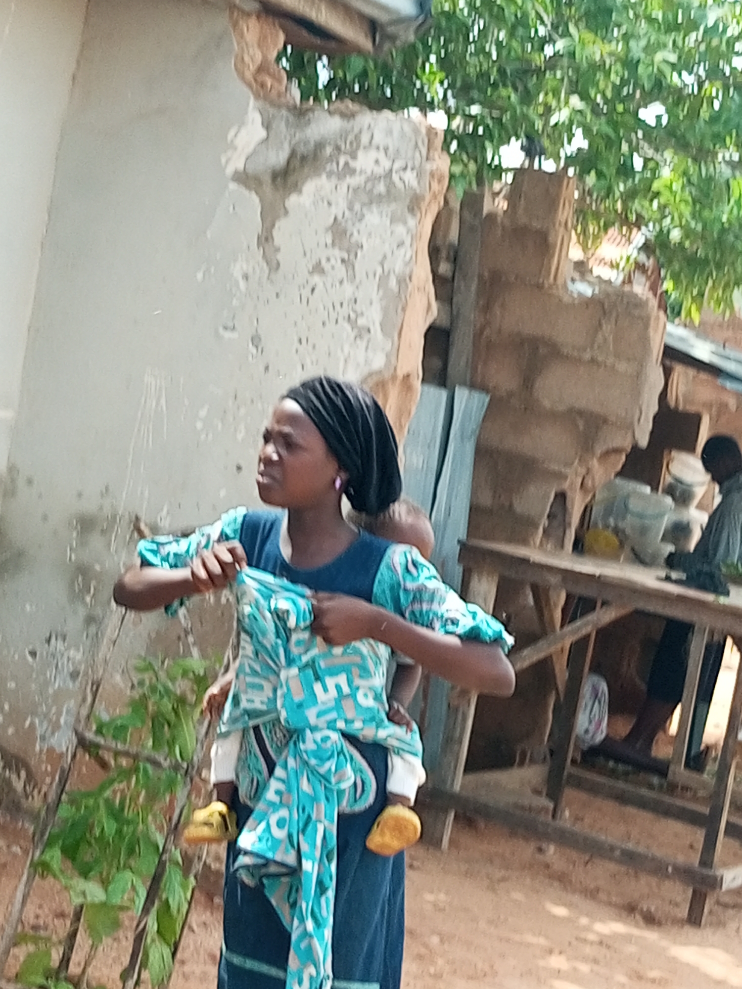 A young woman wearing a black headscarf and a dark blue dress with a turquoise and white patterned wrap is standing and holding a baby on her back. She has a purple earring in her visible ear and yellow shoes on her feet. The baby she is carrying also has yellow shoes. The woman is looking to her left. In the background, there is a damaged building with exposed brick and stucco, a wooden table with items on it, and another person standing behind the table. Trees with green leaves are visible above the structures. The ground is a dusty brown color.