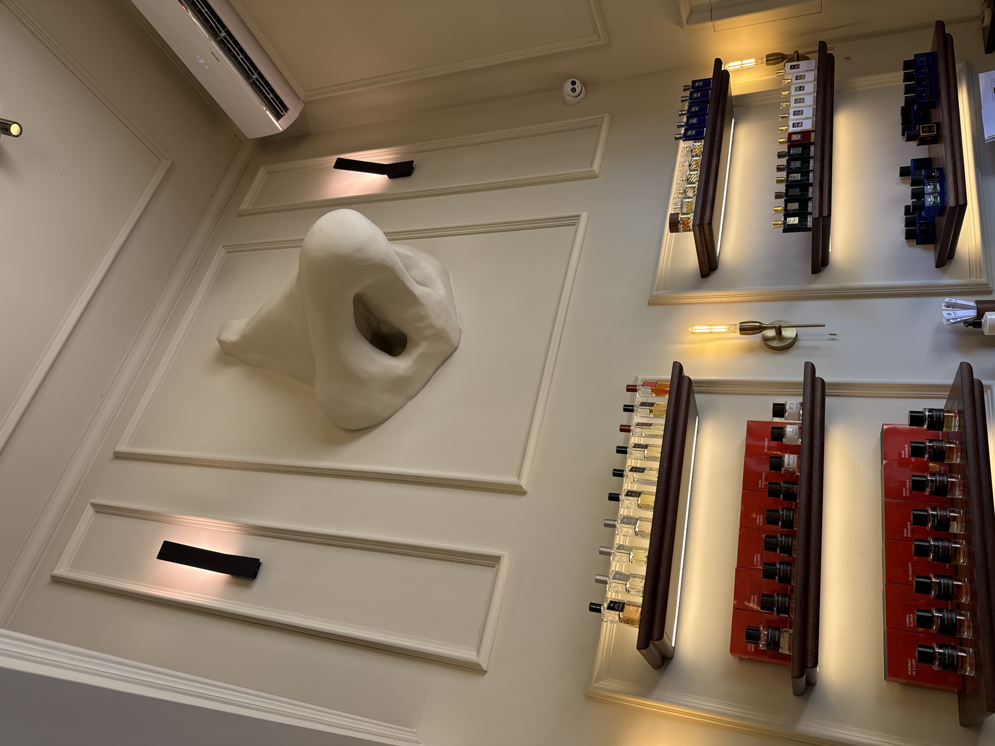 The image depicts the interior of a building in Podgorica, Montenegro. The walls are white with decorative moldings. A large, white sculpture resembling a nose is mounted on the wall. There are several shelves holding bottles of what appear to be fragrances or perfumes. The shelves are illuminated, and the bottles are arranged in neat rows. There are also wall-mounted light fixtures and an air conditioning unit visible in the upper part of the image. No people are visible.