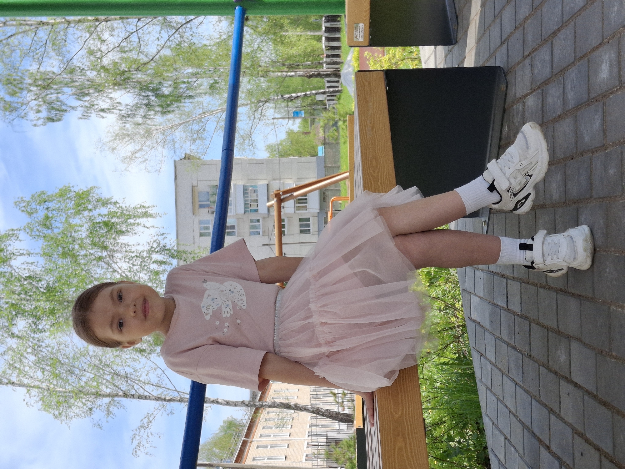 A young girl is seated on a wooden bench in Chaikovsky, Russia. She is wearing a light pink dress with a tulle skirt, white socks, and white sneakers. The background includes a multi-story building, trees, and part of a playground structure with blue and green bars. The surrounding area appears to be a paved walkway with brick-like stones. The lighting suggests it is daytime.