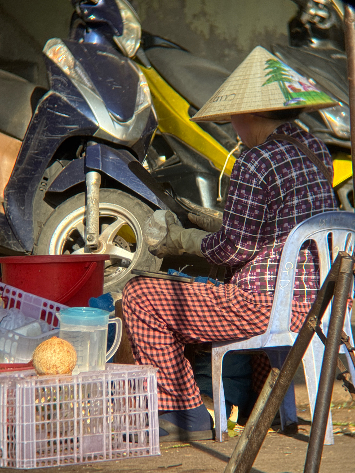 The image depicts a person sitting on a blue plastic chair in what appears to be a street market or outdoor workshop in Nha Trang, Vietnam. The person is wearing a traditional conical hat, a checkered shirt, and checkered pants. They are wearing gloves and appear to be working with their hands, possibly on a coconut or other produce, as indicated by the presence of a coconut and a pitcher of water on a plastic crate in the foreground. A motorcycle is partially visible behind the person, suggesting an urban or semi-urban environment. The lighting suggests it is daytime. The overall scene conveys a sense of everyday life and work in a Vietnamese setting.