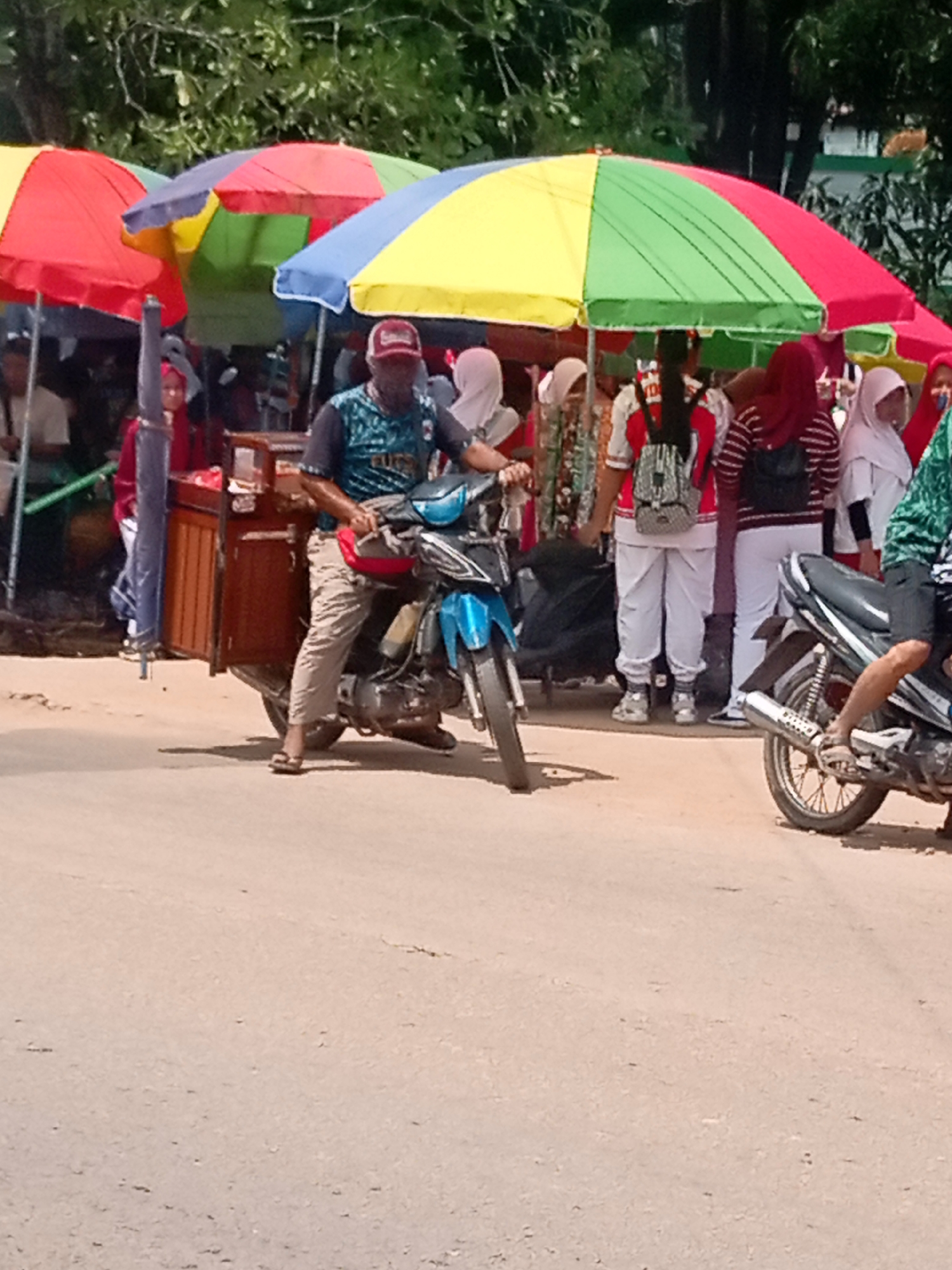 The image depicts an outdoor scene, likely a market or roadside vendor area, in Batam, Indonesia. In the foreground, a man wearing a red cap and a dark blue and teal patterned shirt is riding a blue motorcycle. The motorcycle is equipped with a wooden cart attached to its side, which appears to be a mobile food stall. The cart has a hinged door open, revealing its contents. The man is wearing khaki pants and flip-flops.
Behind the motorcycle, several people are visible, mostly women wearing headscarves. They are standing under a series of colorful, multi-paneled umbrellas. The umbrellas are red, yellow, green, and blue, creating a vibrant canopy. The ground appears to be a dirt or asphalt road. In the background, there are trees and some structures, suggesting an urban or semi-urban environment. Another motorcycle is partially visible on the right side of the image. The overall atmosphere suggests a busy day, possibly during a market day or a street-side gathering.