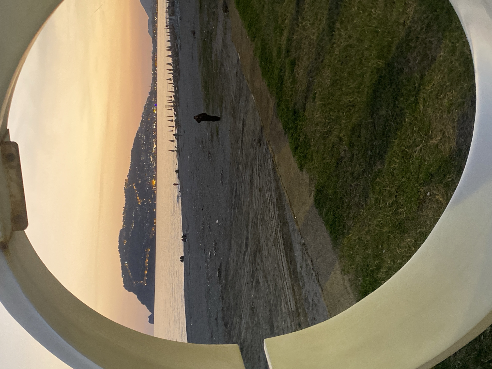 View of Alanya, Turkey at dusk, framed by a circular structure. The scene encompasses a beach, ocean, and a hillside covered with buildings and lights. Beach umbrellas line the shore. A person stands on the beach. Grass and a walkway are to the right of the beach. The sky is a gradient of light orange to yellow.