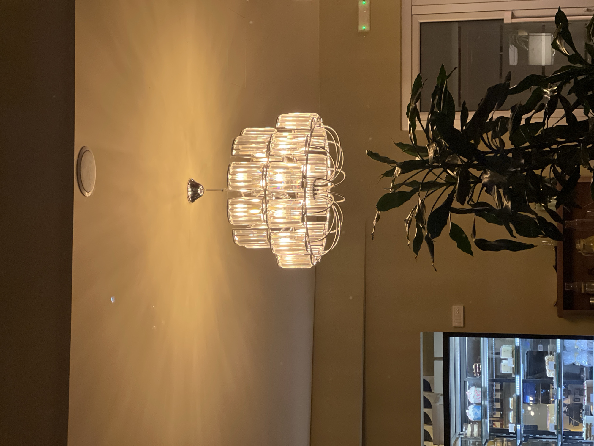 The image shows an indoor scene with a lit chandelier hanging on a beige wall. To the right of the chandelier, there is a window and part of a plant. Below the window is a display case. A circular object is present on the wall to the left of the chandelier. The location is Boreti, Montenegro.