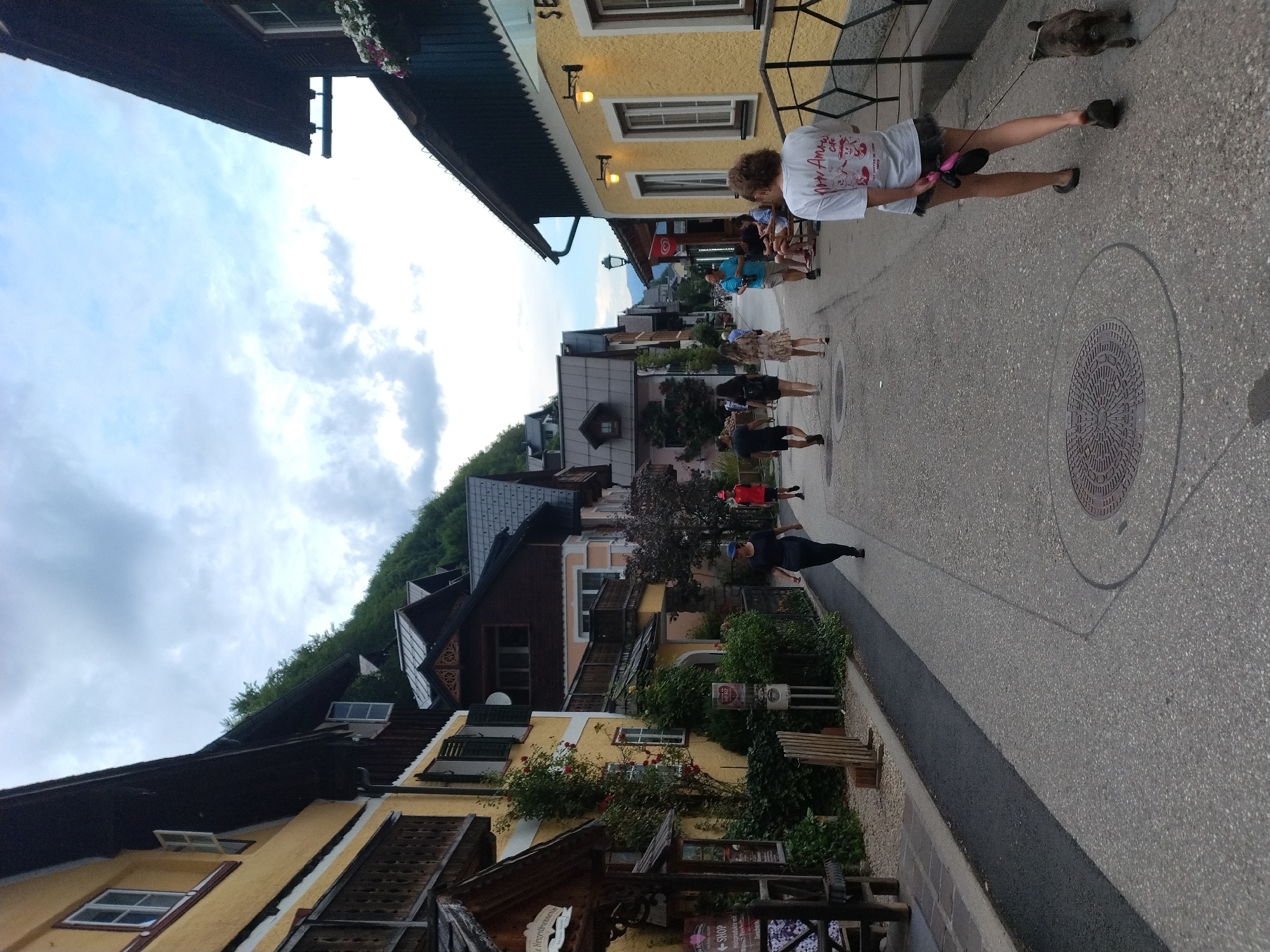 A street scene in Hallstatt, Austria, depicts a narrow, paved walkway lined with traditional buildings. On the left, yellow buildings with dark roofs and balconies display various architectural details, including shuttered windows and ornate trim. Some structures appear to be shops or residences, with signage and awnings visible. Lush green vegetation, including trees and climbing vines, grows along the facades and borders of the walkway. On the right, a yellow building with green trim and exterior lighting is prominent, with a dark railing in front of it.
Several people are walking along the street. In the foreground, a woman with blonde hair walks away from the camera, holding a leash attached to a small, dark-colored dog. She is wearing a white t-shirt with graphics and a dark skirt. Further down the street, a group of individuals are walking towards the camera. One person wears a dark blue shirt and shorts, another a white dress, and others are visible in the distance. A few people are seated on benches outside a building on the right side of the street. The sky overhead is cloudy, with patches of blue visible. The overall impression is a charming, historic street scene in a European town.