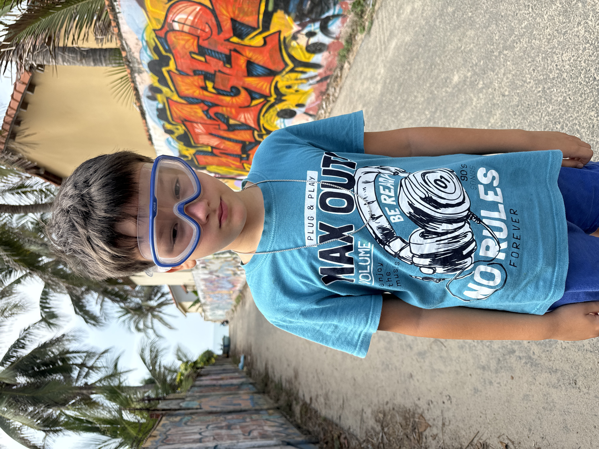 A boy with short, dark hair and blue goggles stands on a paved path in Mũi Né, Vietnam. He wears a blue t-shirt with the text "MAX OUT VOLUME PLUG & PLAY" printed on it and a chain necklace. Graffiti art is visible on a wall behind him. Palm trees and other vegetation are present in the surrounding environment.