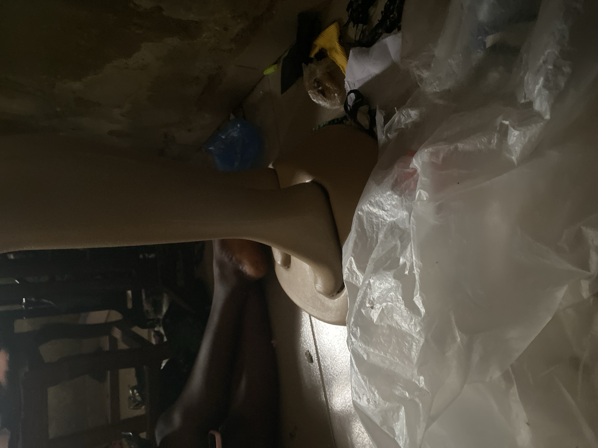 An indoor scene depicts a close-up of a mannequin leg and foot, made of pale material, standing on a tan-colored base. To the left, a dark-skinned person's leg and foot are visible, resting on the floor. The environment appears to be cluttered, with various objects scattered around, including white plastic sheeting in the foreground, a blue plastic bag, a yellow object, and dark debris. The floor appears to be tiled. The background suggests a dimly lit area with wooden structures and a textured wall. The scene is located in Jauro Yinu, Nigeria.