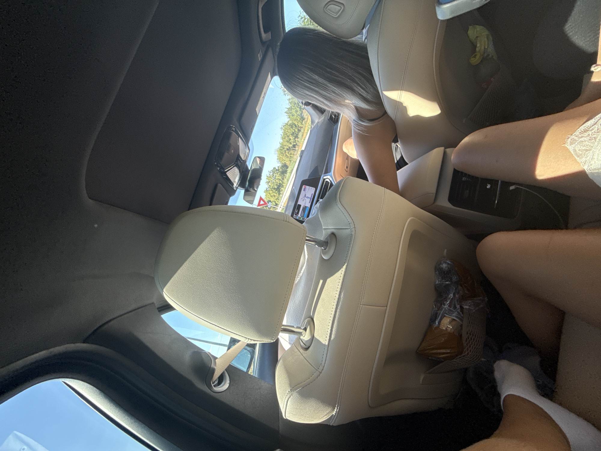 The image shows the interior of a car, viewed from the back seat. A person with blonde hair is in the driver's seat, with their back to the camera. They are wearing a black top. The car is driving on a sunny day, with trees visible outside the windows. A navigation screen is visible on the dashboard. The seats are leather and a light beige color. In the back seat, a person's legs are visible, wearing white socks and a white lace garment. A brown plastic-wrapped object is in a mesh pocket on the back of the driver's seat. The image does not provide any specific information about the location in the city of Simila, Romania, or any other specific geographic location.