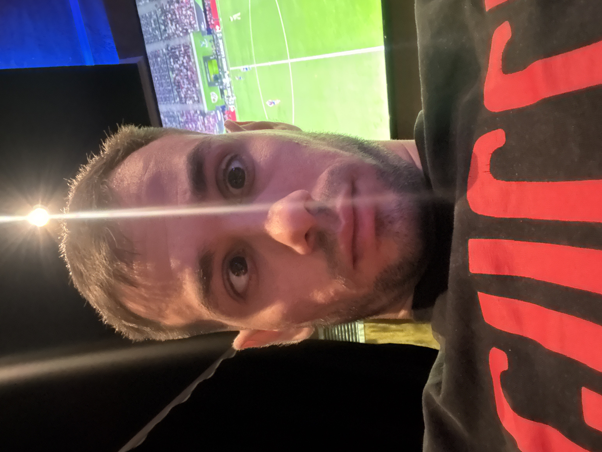 A close-up, rotated image depicts a light-skinned male with short hair, a mustache, and a beard stubble. He is wearing a dark shirt with large red markings. The background includes a television screen showing a soccer match and a dark, possibly outdoor setting. The image is likely taken indoors. There is no indication of the location in Burgas, Bulgaria.