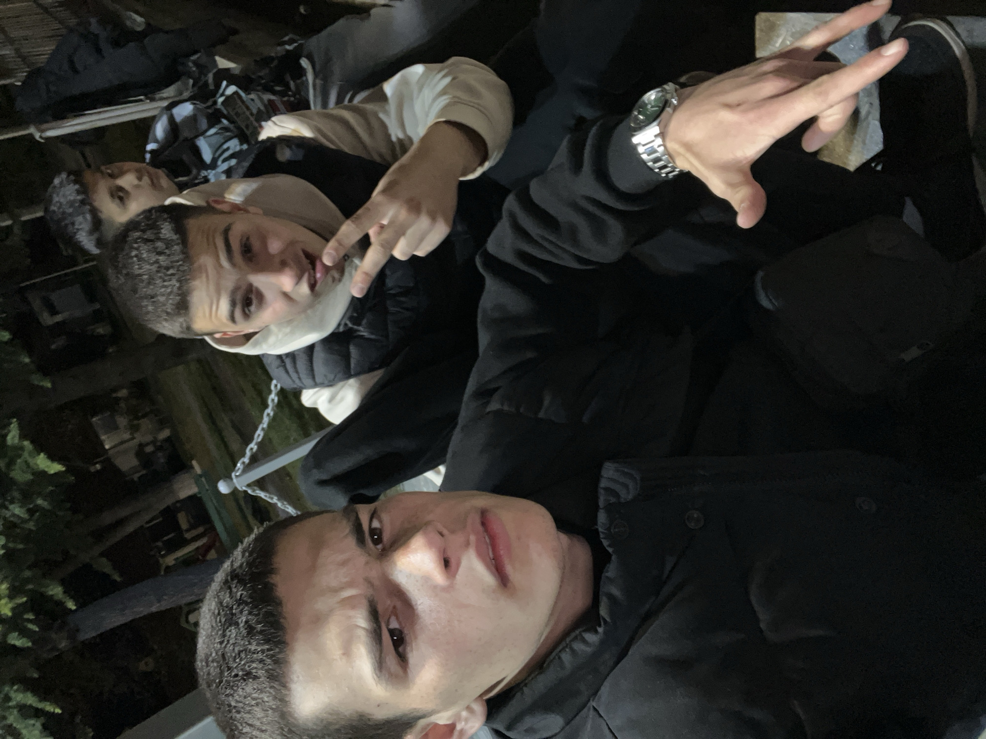 Three young men are seated outdoors in Karavelovo, Bulgaria. The man in the foreground has dark hair and is wearing a black jacket, looking directly at the camera with a slight frown. Behind him and slightly to the left, another man is making a peace sign with his fingers, wearing a white hoodie under a black vest. Further behind, a third man is partially visible, also with dark hair. The environment is dimly lit, and details of the surroundings are limited, though there appears to be some form of metal structure with a chain visible in the background. The closest man is also wearing a watch.