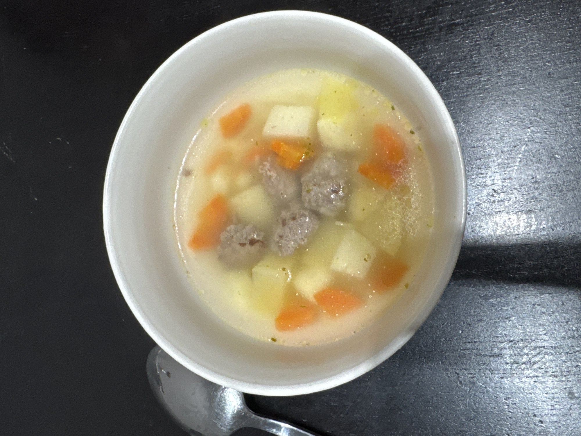 A top-down view of a white bowl filled with soup, likely on a dark, possibly wooden, surface. The soup contains broth, cubed potatoes, sliced carrots, and several small meatballs. A metal spoon rests near the bottom left of the bowl.
The location in the city of Perm, Russia, is not identifiable from this image.