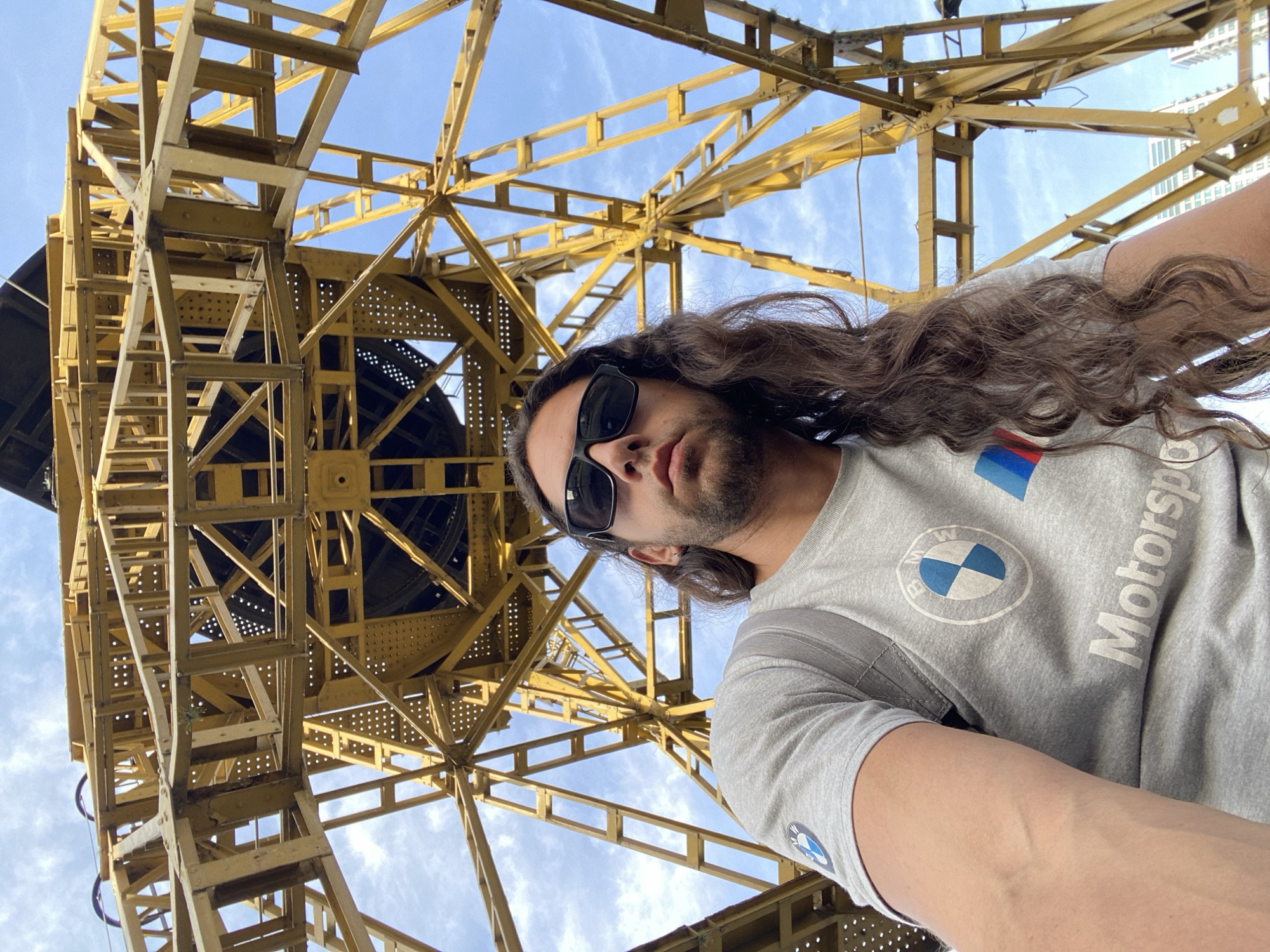 A man with long brown hair and sunglasses is taking a selfie. He is wearing a gray BMW Motorsport shirt. Behind him is a large yellow metal structure against a blue sky with some clouds. The structure appears to be the Torre Espacial, a communications tower located in Buenos Aires, Argentina.