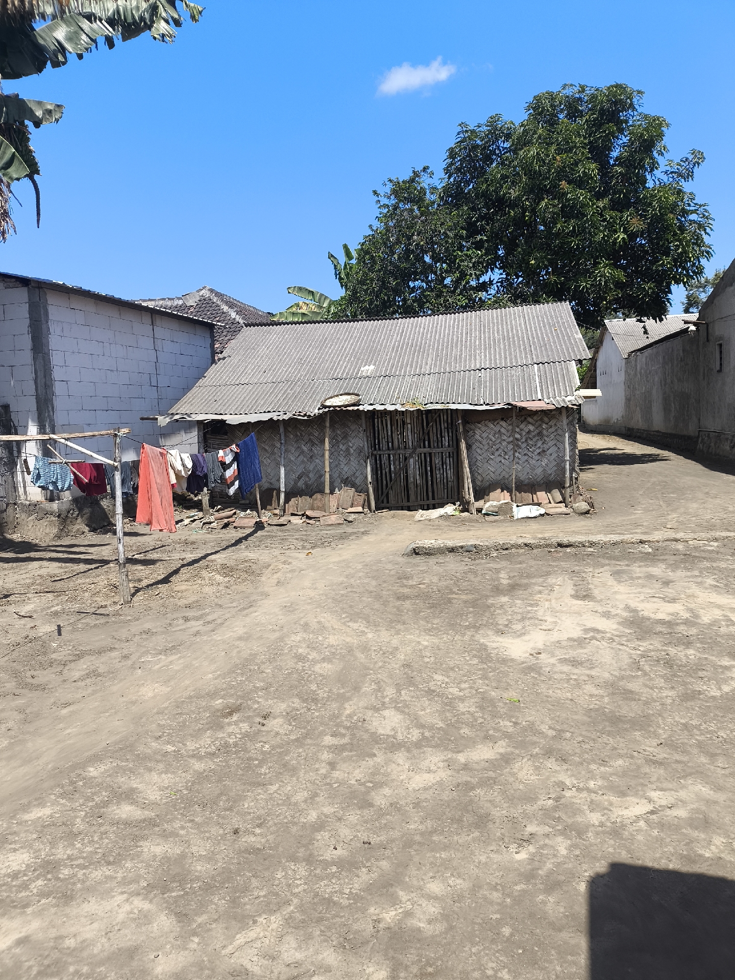 The image depicts a scene in the city of Grati, Indonesia, on a bright, sunny day. In the foreground, a dusty, unpaved ground stretches out, marked with tire tracks and some scattered debris. A clothesline with various garments, including red, blue, and striped shirts, is strung between two wooden poles in front of a simple, single-story structure. This building features a corrugated metal roof and walls constructed from woven bamboo. Adjacent to this structure is a larger building with white brick walls. Further in the background, more buildings with similar roofs can be seen, and a large, leafy green tree stands tall against a clear blue sky with a few wispy clouds. The overall impression is of a rural or semi-rural residential area.