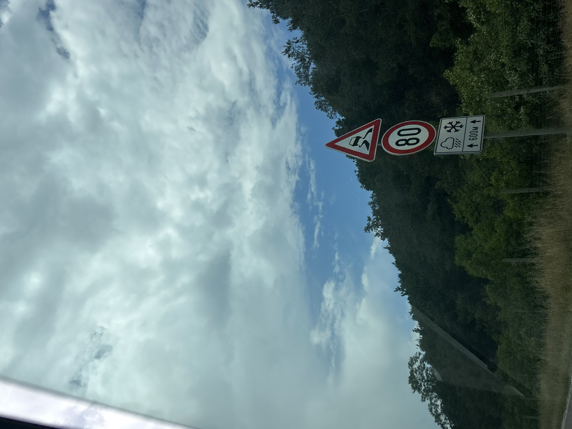 The image depicts a rural mountainous scene in Gorno Varshilo, Bulgaria, viewed from inside a vehicle, evidenced by a glare on the left. The upper two-thirds of the frame features a partly cloudy sky with patches of blue visible amidst greyish-white clouds. Below the sky, a dense, dark green forested hillside slopes from left to right across the mid-ground. A tall, multi-component road sign stands on the right side, against the backdrop of the forest. The topmost sign is a red-bordered white triangle with a black skidding car symbol, indicating a slippery road. Below it is a red-bordered white circular sign displaying "80" in black, signifying a speed limit of 80. The bottom sign is a white rectangle with symbols for rain and snow, and the text "↑ 600M ↑", likely indicating a distance for the depicted conditions. The immediate foreground shows dry, light brown grass and sparse green vegetation along what appears to be a roadside. No individuals or active subjects are visible.