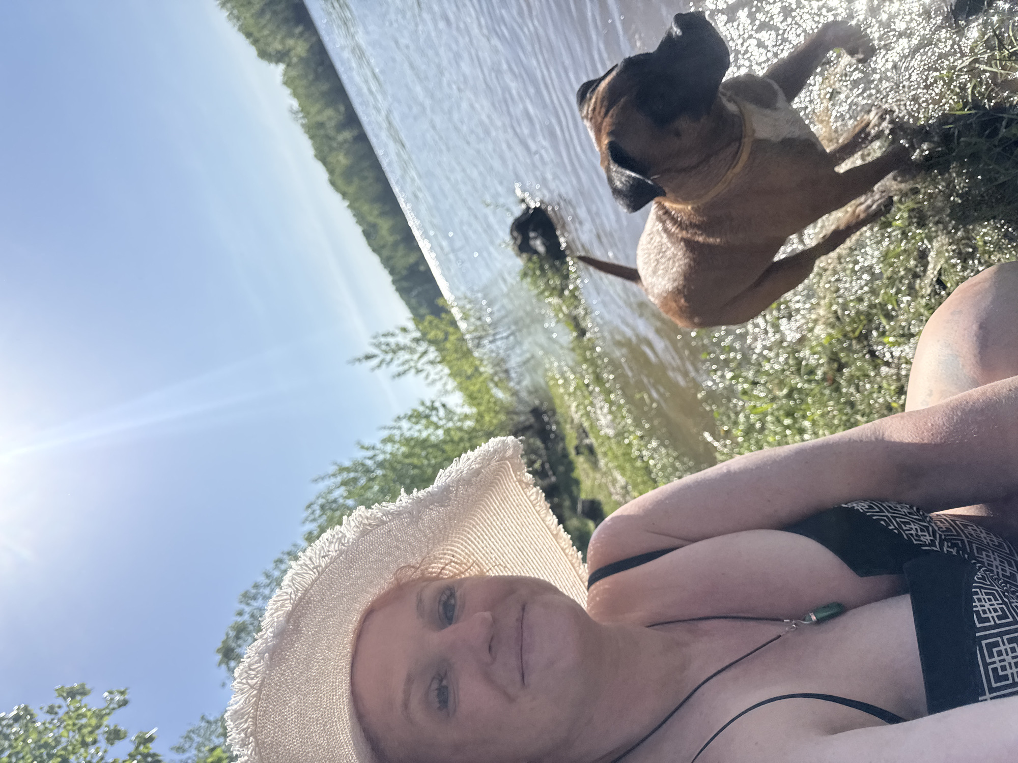 A woman wearing a straw hat and a black swimsuit is lying near a body of water. Two dogs are present; one is near the shoreline and the other is farther out in the water. The background includes greenery and trees, suggesting a natural setting. This is in the city of Chaikovsky, Russia.