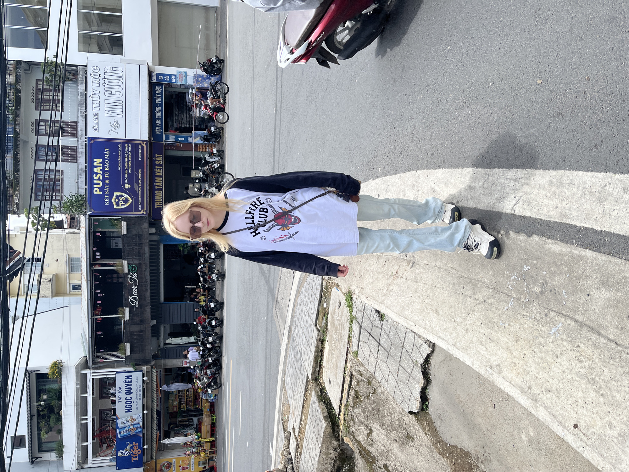 A young Caucasian woman with blonde hair and sunglasses walks on a sidewalk in Da Lat, Vietnam. She is wearing a white long-sleeved shirt with black sleeves, featuring a graphic design of a red demon with "HELLFIRE CLUB" written above it. She is also wearing light blue jeans and white and black sneakers. A brown crossbody bag is slung over her shoulder.
The street scene in the background shows several buildings with signage in Vietnamese. One building has a sign that reads "TẬP HÓA NGỌC QUYÊN" with "Tiger" branding. Another building has signs for "PUSAN KẾT SẮT & TỦ BẢO MẬT" and "KIM CƯỜNG". The sidewalk is made of concrete slabs, some of which are broken. Numerous motorbikes are parked along the street, and a few are in motion. The weather appears to be sunny, casting shadows on the pavement. The environment suggests an urban setting with commercial establishments.