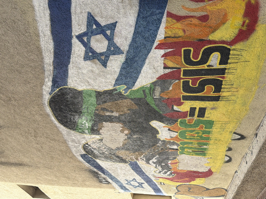 A mural is displayed on a light beige, textured stucco wall in Tel-Aviv, Israel. The central element of the mural is a large depiction of the Israeli flag, featuring two horizontal blue stripes on a white background with a central blue Star of David. Overlaid onto the flag are two stylized, dark-colored figures, each wearing a green headband bearing the Arabic script "حماس" (Hamas). The face area of the leftmost figure shows significant wear or deliberate obscuring, revealing the underlying wall surface. Below these figures, large, stylized graffiti reads "MAS = ISIS," with "MAS" in green dripping letters and "= ISIS" in black dripping letters. Red and orange flame motifs emanate from behind and around this text. The lower portion of the wall exhibits faint, indistinct black graffiti. A small, dark-recessed window is visible in the upper left corner of the building. The scene is illuminated by direct sunlight.