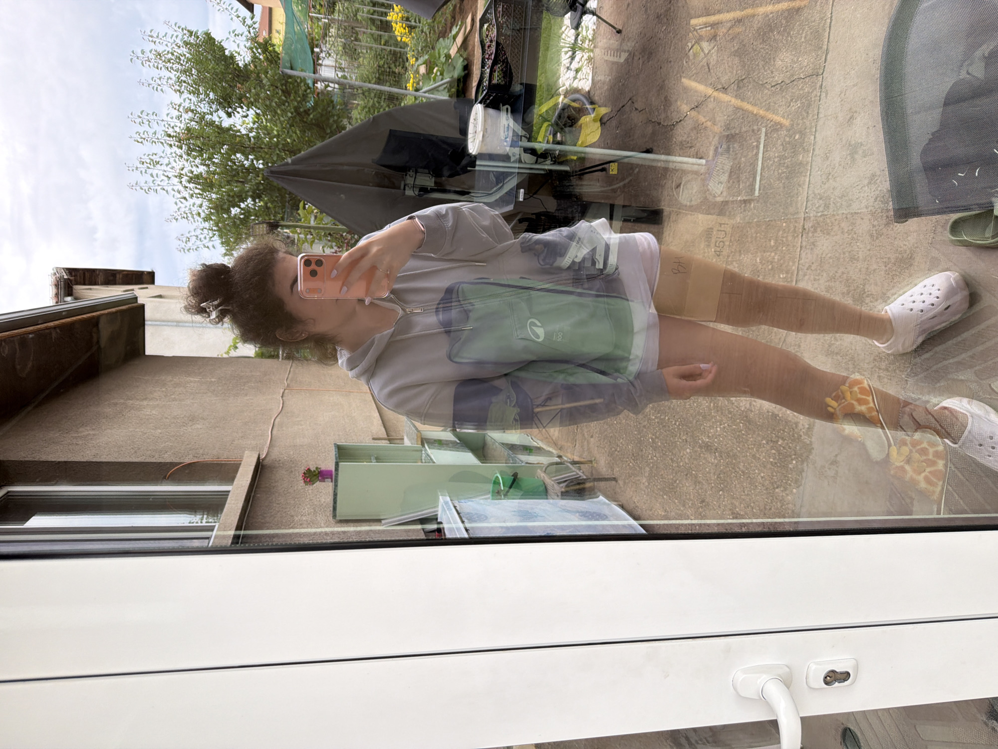 A young woman with curly brown hair styled in a top knot is taking a mirror selfie in what appears to be a backyard or patio area. She is wearing a light gray zip-up hoodie over white shorts and white clogs. The hoodie has a large green graphic on the front. She is holding a pink smartphone up to take the photo.
The background, reflected in the glass, shows a residential building with a textured beige exterior and a tiled roof. There is a window on the side of the building. In the yard, there is outdoor furniture, gardening supplies, a tent-like structure, and lush greenery with yellow flowers. The ground is paved with concrete. A white door with a silver handle and lock is prominent in the foreground on the left side of the image, suggesting the selfie is taken through a glass door or window of a residence. A brown band with writing is visible on the woman's right thigh. The image is likely taken in Sofia, Bulgaria, based on the context provided.