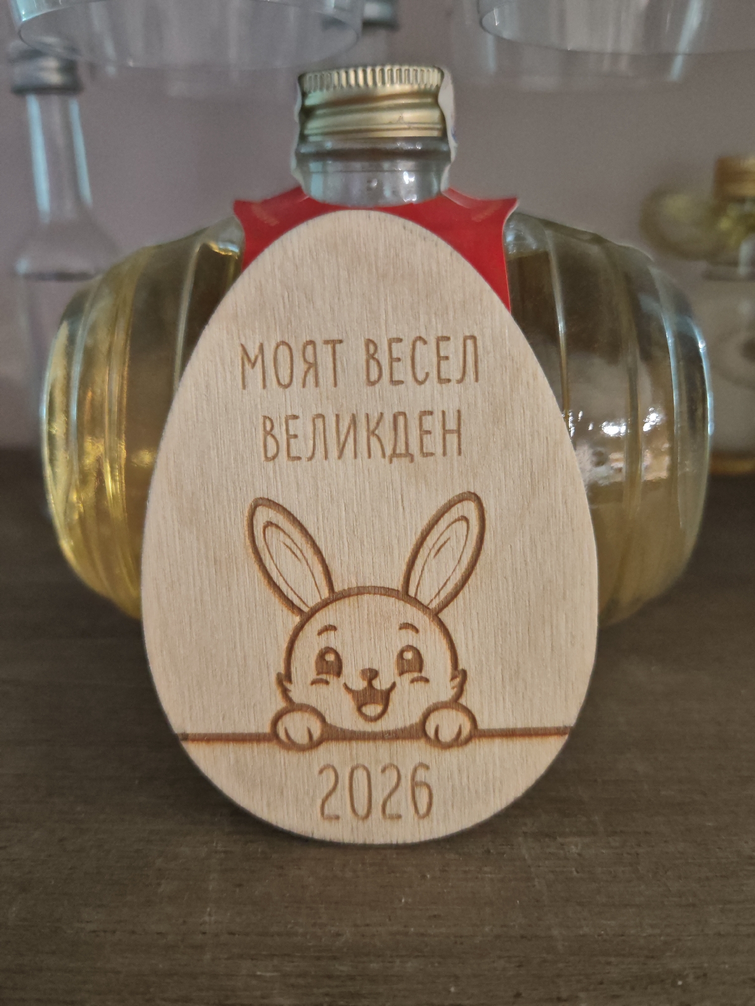 A close-up, eye-level shot shows a wooden egg-shaped ornament placed in front of a glass bottle with a ribbed texture and yellow liquid. The ornament features a cartoon drawing of a smiling rabbit with big ears and paws peeking over a horizontal line. Above the rabbit, the ornament is inscribed with the Cyrillic text "МОЯТ ВЕСЕЛ ВЕЛИКДЕН," which translates to "My Happy Easter." Below the rabbit and the line, the year "2026" is engraved. A small red ribbon is affixed to the top of the ornament. The background is blurred, showing the tops of other glass bottles and a grey, textured surface. The ornament appears to be a souvenir or decoration. The image does not contain any identifiable landmarks or clues to suggest a specific location in Plovdiv, Bulgaria.