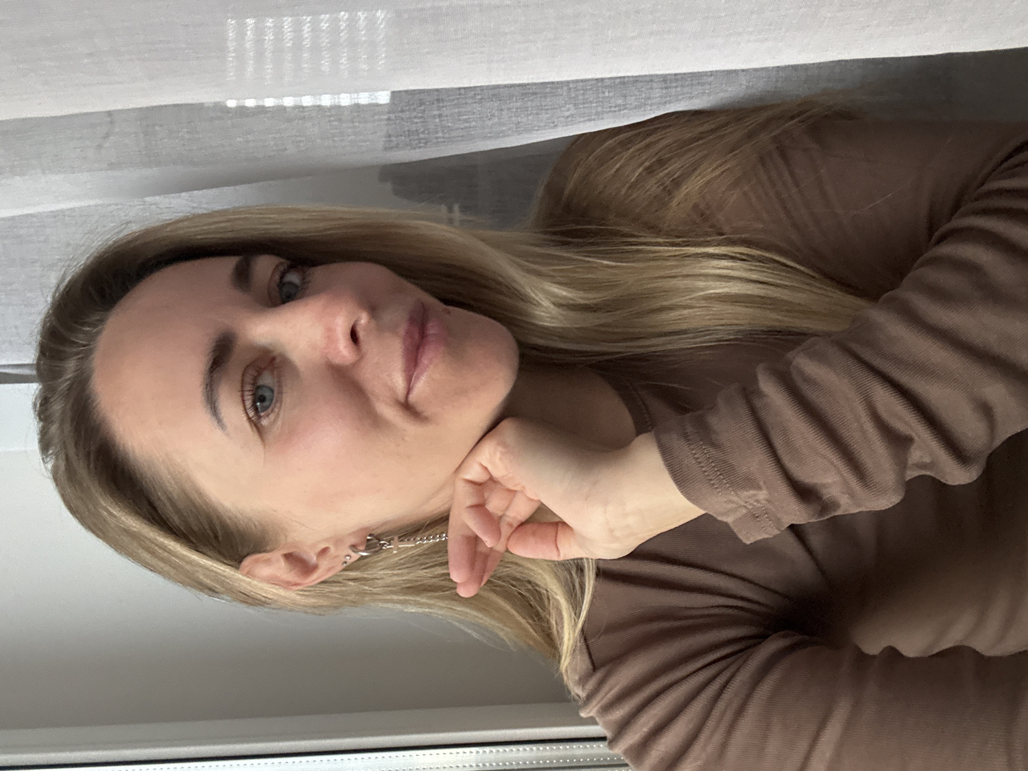 A head-and-shoulders portrait features a young woman with long, straight blonde hair and blue eyes. She is looking directly at the camera with a gentle, slightly closed-lip smile. Her right hand is raised, with her fingers loosely resting near her jawline and chin. She wears a long-sleeved, ribbed top in a warm brown or taupe color. In her left ear, she has a silver chain-style earring and multiple small hoop piercings.
The setting appears to be indoors, with a soft, natural light illuminating her face, suggesting daytime. A white or light-colored curtain or blind is partially visible in the upper background, indicating she is positioned near a window. The lower part of a window frame or sill is also visible. The overall mood is calm and friendly. No specific location cues within the image point to Chaikovsky, Russia, beyond it being an indoor portrait. There is no visible text.