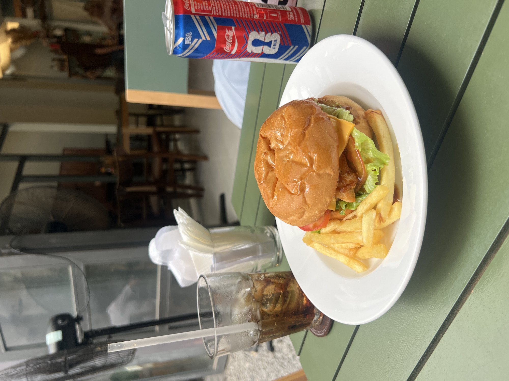 The image shows a meal consisting of a burger and fries served on a white plate, next to a Coca-Cola can and a glass of iced soda with a straw. A container of tissues sits nearby. The scene is set on a green, likely outdoor table. The setting is Bac Nha Trang Ward, Vietnam, based on the soda can that is labeled "FRA", indicating it is made in Nha Trang. The surrounding environment is visible in the background, showing a blurred restaurant interior with tables and chairs. There is no indication of human presence in the immediate foreground, only the served food and beverages.
