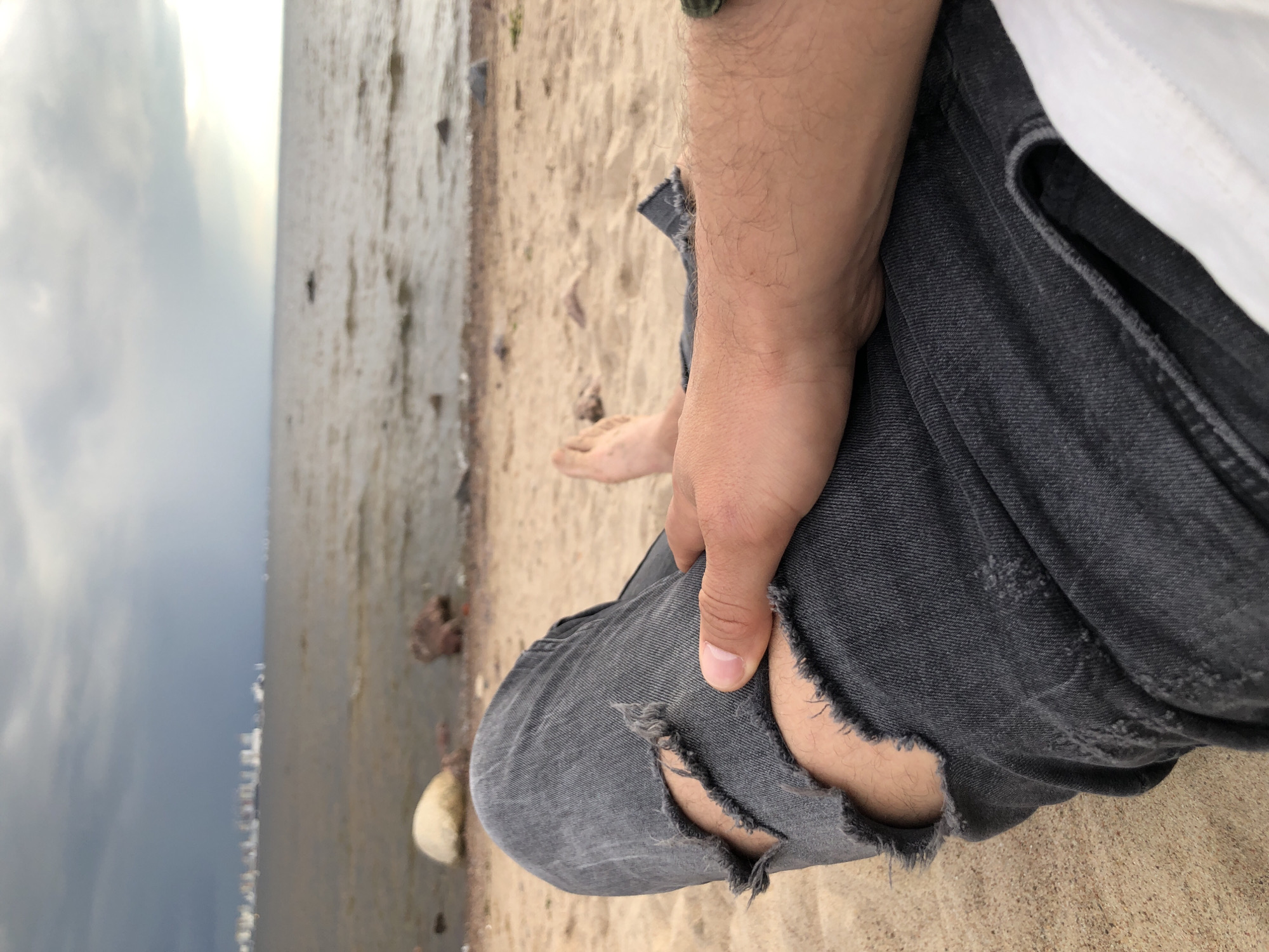 A first-person perspective shot of a person relaxing on a sandy beach near a body of water. The person is wearing ripped charcoal grey jeans and a white shirt, and their hand rests on their thigh. The background shows a calm body of water with a distant cityscape under a cloudy sky. The weather appears overcast, possibly indicating an approaching storm.