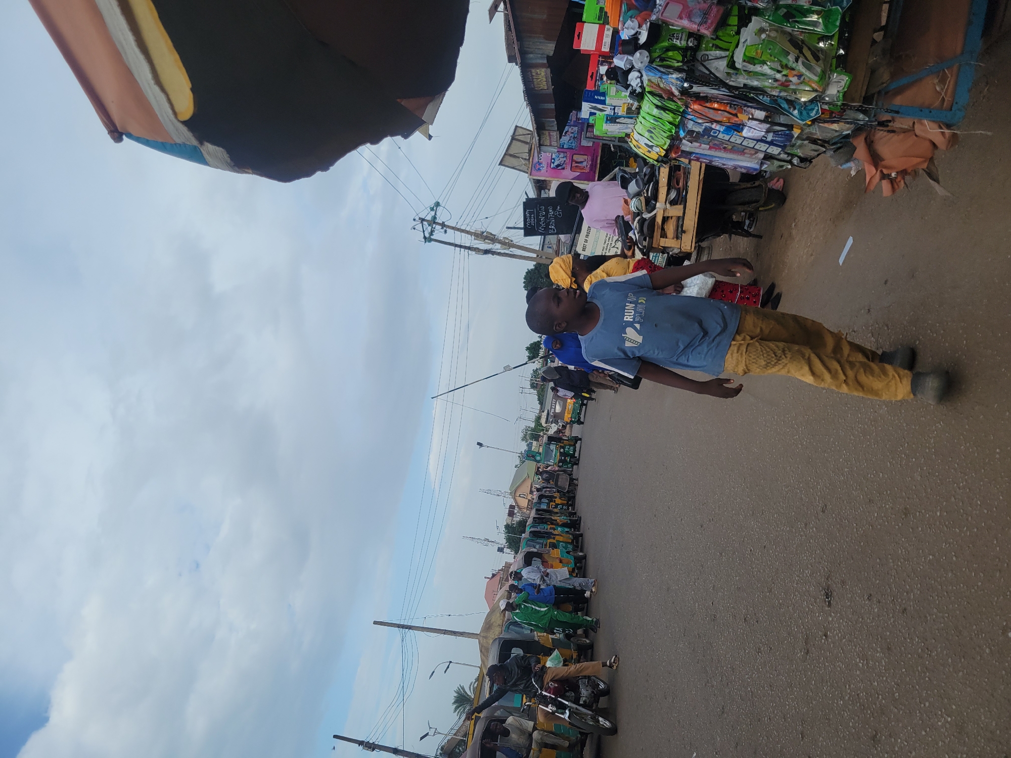 The image captures a lively street scene in Jos, Nigeria, under an overcast sky. The photograph is taken from a slightly elevated and tilted perspective, looking down a bustling road flanked by commercial activity.
In the foreground, a young individual, likely a boy, is prominent, dressed in a blue t-shirt emblazoned with "RUN UP PLAY" and mustard yellow trousers. He stands on the asphalt road, looking towards the left side of the frame with his right arm slightly raised.
To the right, a series of market stalls display a wide array of goods, including various packaged items, footwear, and electronics. A person in a light pink shirt is visible near one of these stalls. A black signboard, hanging from a stall, presents white text that includes phrases like "MAMMY PURE," "AKWAKAWA," "BUNI FOOD," and "BEST OF FRUITS."
Along the left side of the street, a long queue of green and yellow tricycles, known locally as Keke Napep, are parked, suggesting a transport hub or waiting area. Numerous people are seen interacting with these tricycles, some seated, others standing. Further down the road, a person is also seen on a motorcycle. Utility poles with a web of overhead wires are visible, typical of an urban setting.
The scene depicts everyday life in a busy urban environment, possibly a market or commercial district, characterized by a mix of vendors, commuters, and pedestrians. The cloudy sky suggests it is not mid-day with bright sunshine.