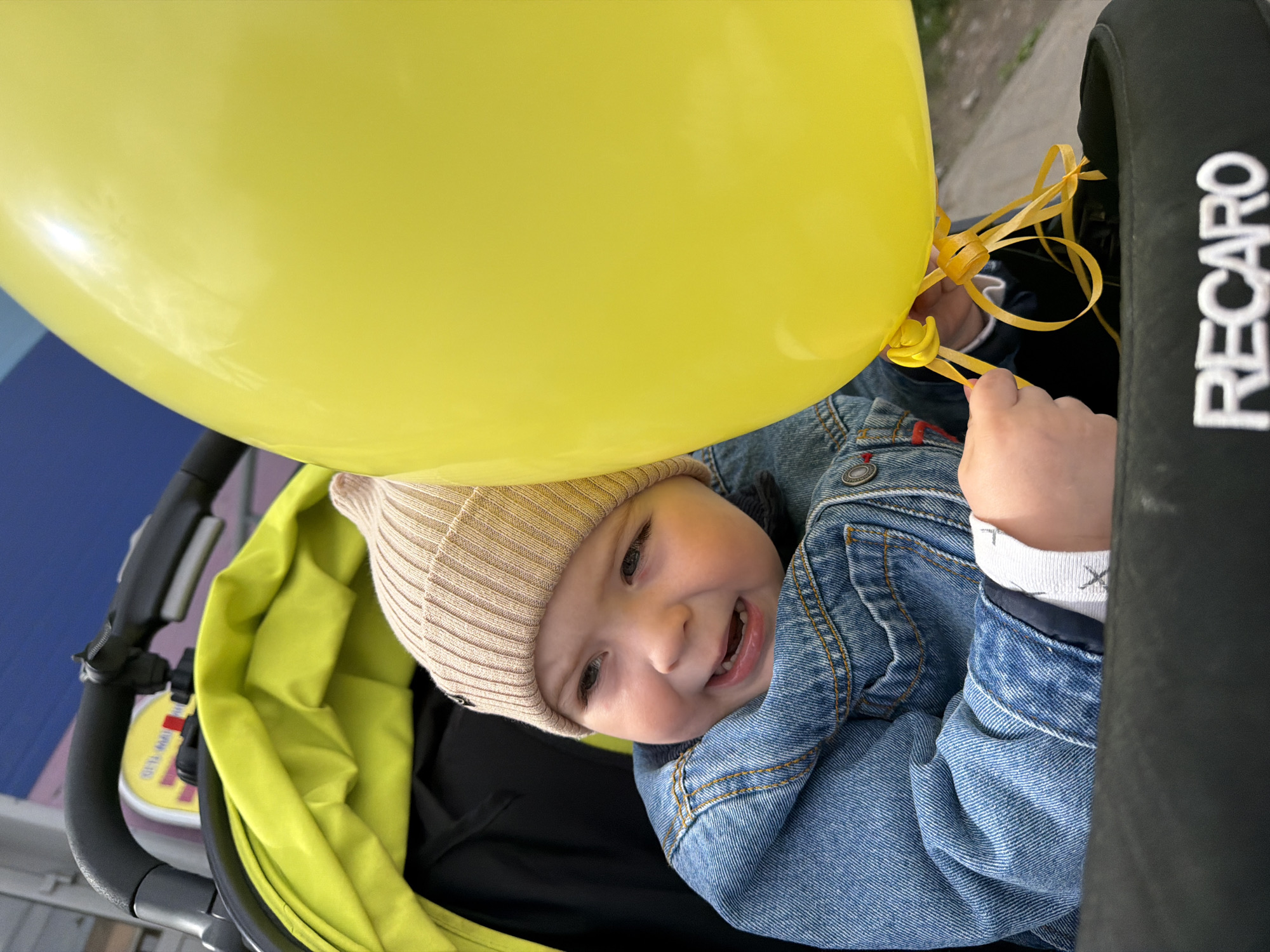 A baby wearing a tan beanie and a denim jacket sits in a bright green stroller. A large yellow balloon is partially obscuring the baby's face. The baby is smiling and reaching out to hold the balloon strings. The side of the stroller reads "RECARO" in white letters. The location cannot be confirmed as Votkinsk, Russia based on visual information.