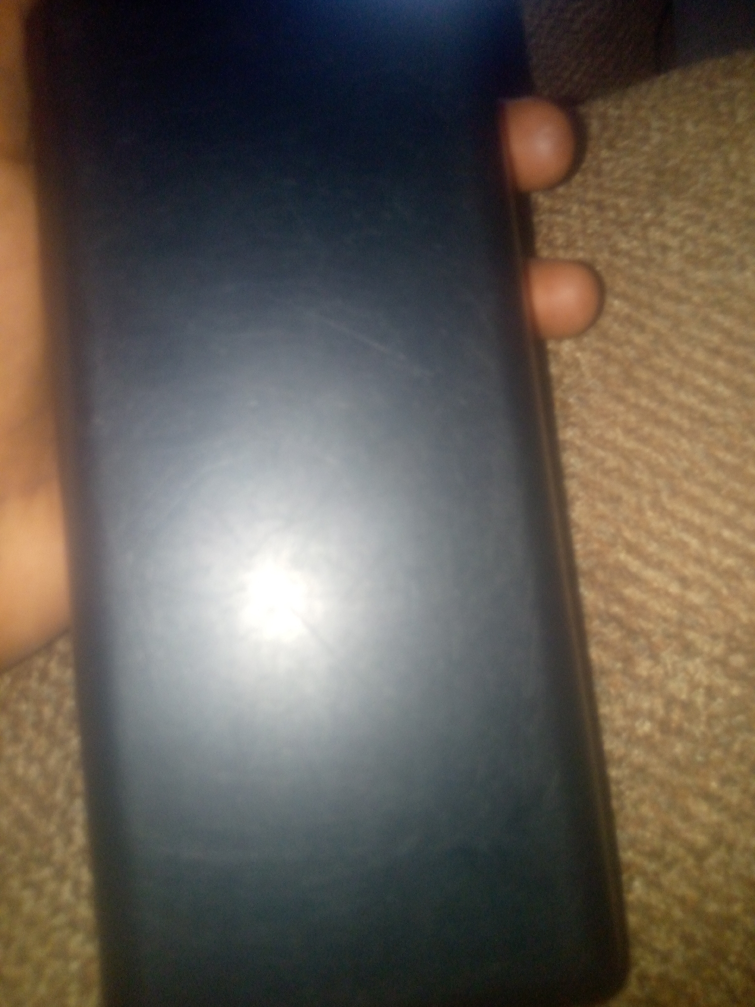 The image shows a dark blue rectangular object, possibly a phone or a power bank, lying on a brown, textured surface resembling a woven blanket or upholstery. A person's hand is visible on the left side of the image, holding the object. Two fingers, likely from the same hand, are visible on the right side, pressing against the brown surface. The object has a matte finish and appears to be smooth, with a bright light reflection on its surface. The overall lighting suggests an indoor setting. The image does not contain any information about the city of Jauro Yinu, Nigeria.