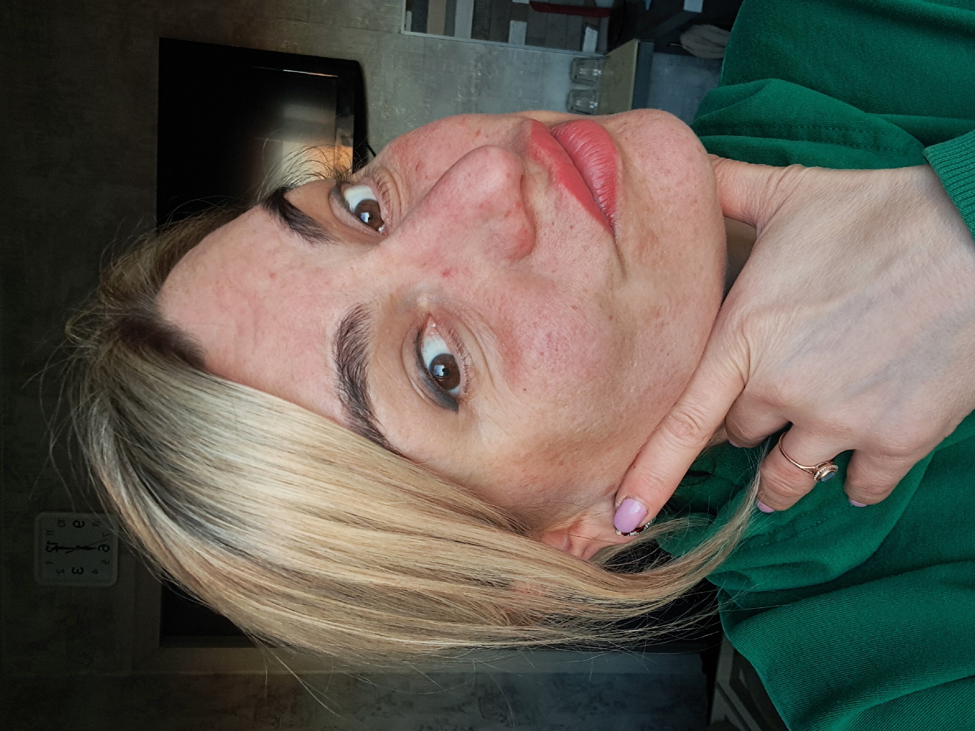 A woman is facing the camera at an angle, wearing a green sweater. Her right hand touches her face. She has blonde hair and is wearing lipstick. A television is visible behind her. A clock reads 5:12. The location is Elektrostal, Russia.