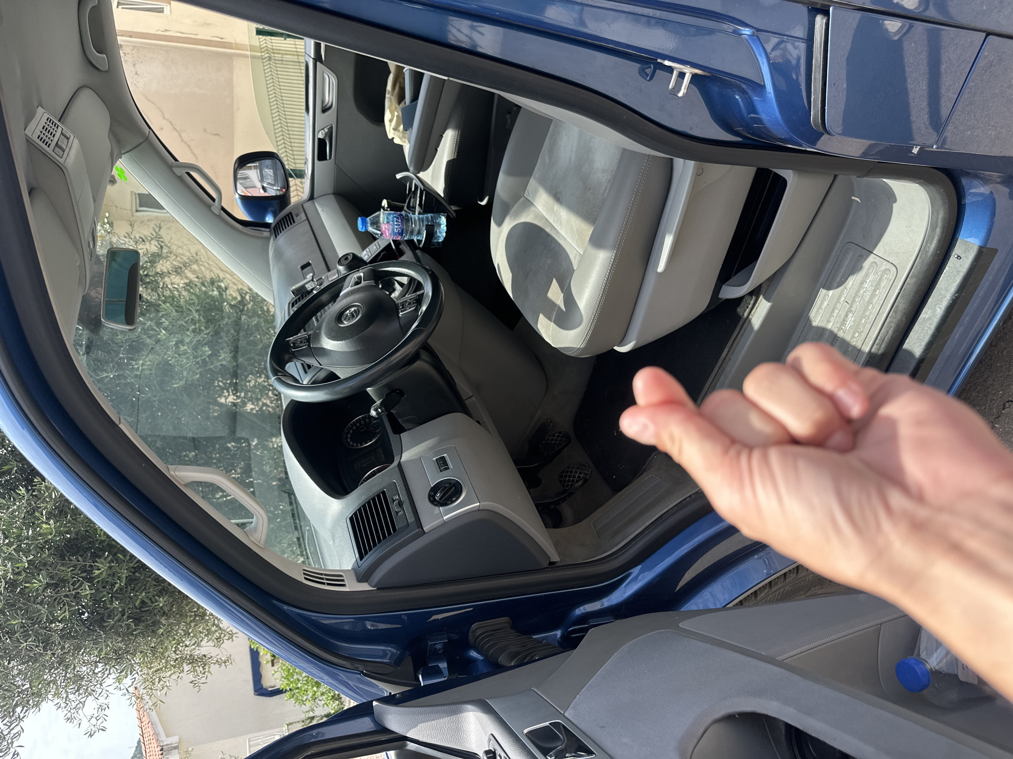 This media file captures the interior of a blue Volkswagen vehicle, likely a van or SUV, viewed from the open driver's side door. The setting is outdoors in Trap, Montenegro, under bright, sunny daytime conditions, evidenced by clear light and shadows within the vehicle.
A light-skinned hand, presumably the photographer's, is prominent in the foreground, partially obscuring the lower right of the image. The vehicle's interior features light grey or beige cloth seats, a dark grey dashboard, and a steering wheel bearing the distinctive "VW" (Volkswagen) logo. Visible dashboard elements include air vents, a rotary light switch, and climate controls. On the center console or between the seats, a transparent plastic water bottle with a blue cap is visible, clearly labeled with the brand name "SUZANA".
Through the windshield and driver's window, a background of a light-colored building and green foliage (trees or bushes) can be seen, reflecting the local environment. An exterior side mirror is visible, showing a reflection of the surroundings. An overhead console with various buttons is also present above the windshield.
The activity depicted is a static view of the car's interior with the door open, implying a moment of access or documentation. No other individuals are present in the scene. The overall impression is of a clean vehicle interior situated in an outdoor, possibly residential or semi-urban, sunny Mediterranean environment.