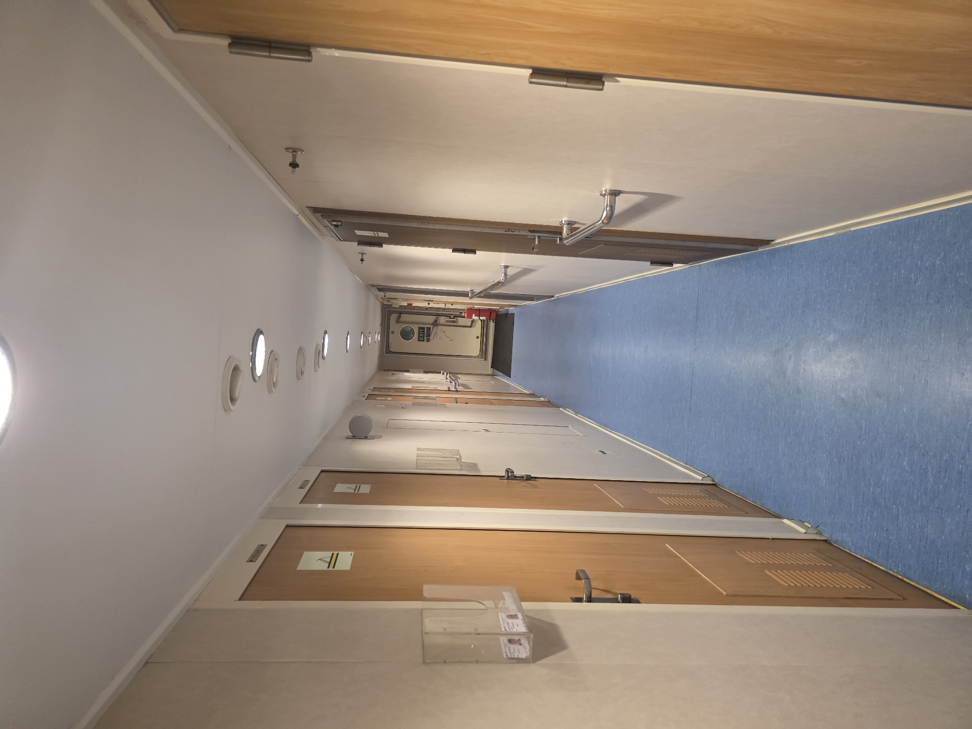 The image depicts a long, narrow corridor with doors on either side. The corridor has a blue floor and a white ceiling with circular lights. The walls are a light beige color. On the left side, there are several wooden doors with metal handles and signs above them. There are also clear plastic holders with what appear to be identification cards attached to the wall next to some doors. On the right side, there are also wooden doors and metal handrails along the wall. Towards the end of the corridor, there is a door with an "EXIT" sign and a red fire extinguisher next to it. The overall impression is of a functional interior space, possibly on a ship or in a building with many rooms. There is no information in the image that would suggest a specific location in Brindisi, Italy.