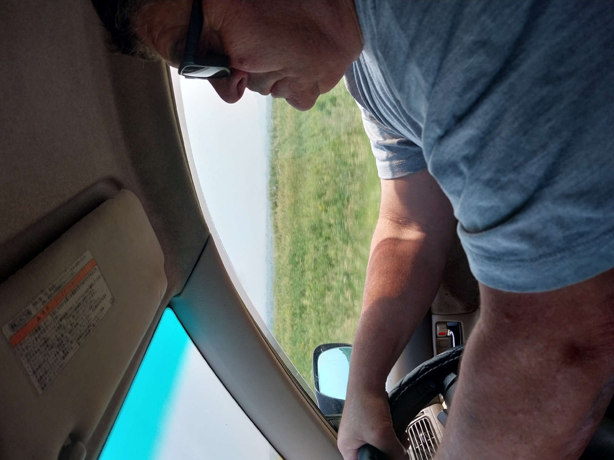A medium shot captures a person driving a car, with the camera positioned inside the vehicle and looking forward. The driver, wearing a grey t-shirt and sunglasses, is holding the steering wheel with their right hand. Their left arm is visible resting on the steering wheel. The interior of the car is visible, including the dashboard, a side mirror, and a sun visor with Japanese text. Outside the car, a blurry green landscape with a hazy sky can be seen through the windshield and side window. The image does not provide any specific details about the city of Sychevka, Russia.