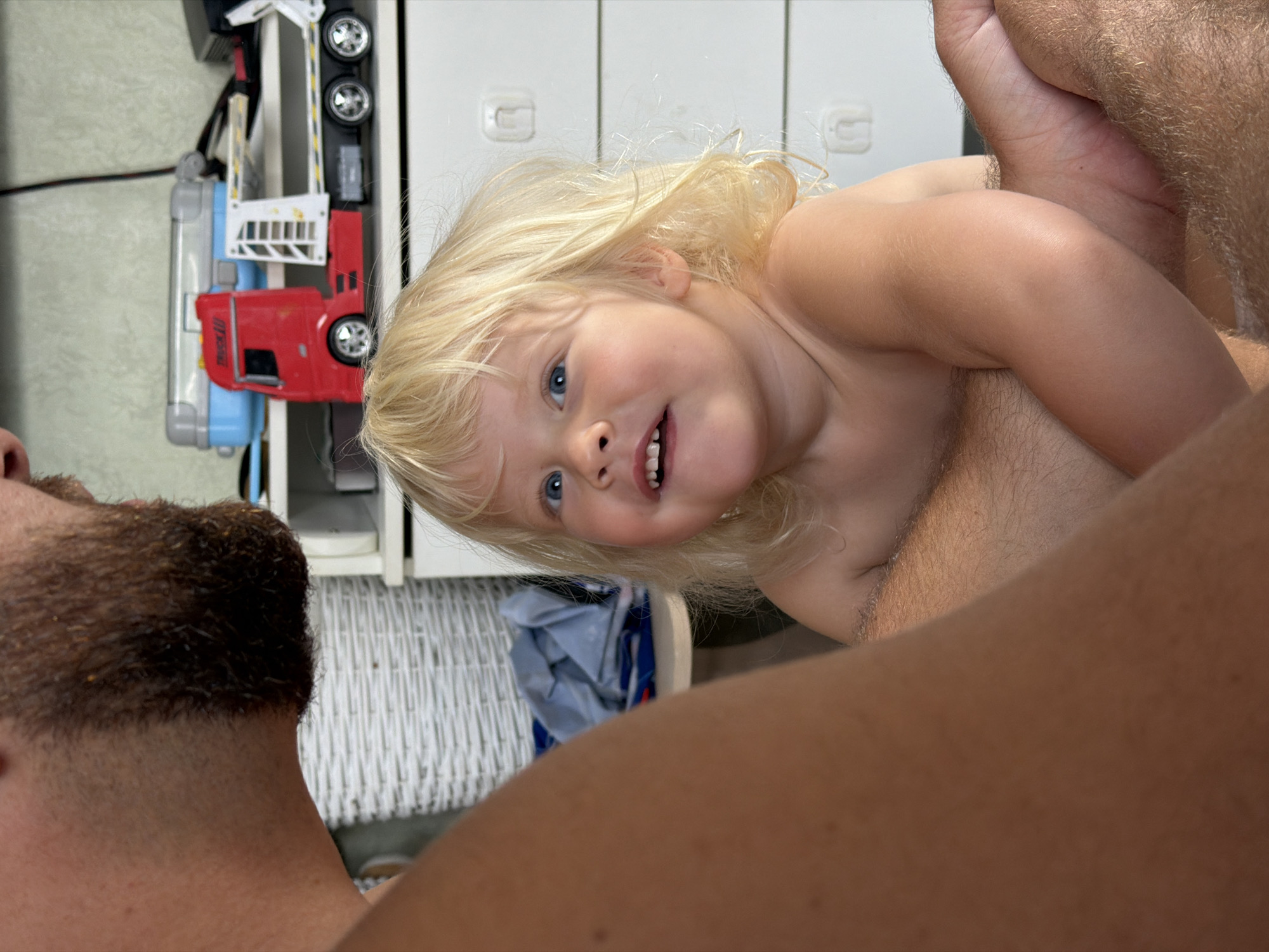 This media file features a close-up, slightly angled shot of a toddler and an adult. The toddler, with blonde hair and blue eyes, is looking towards the camera with a playful expression. The adult's face is partially obscured, but their arm and shoulder are visible. They appear to be holding the toddler.
In the background, a toy truck is visible, with red, blue, and silver components, and wheels. It is positioned on a white surface that resembles shelving or storage units. The overall setting appears to be indoors, possibly a room with white furniture and a woven basket in the background. The lighting suggests it is daytime. There is no discernible text in the image. The context provided indicates the location is Sochi, Russia.