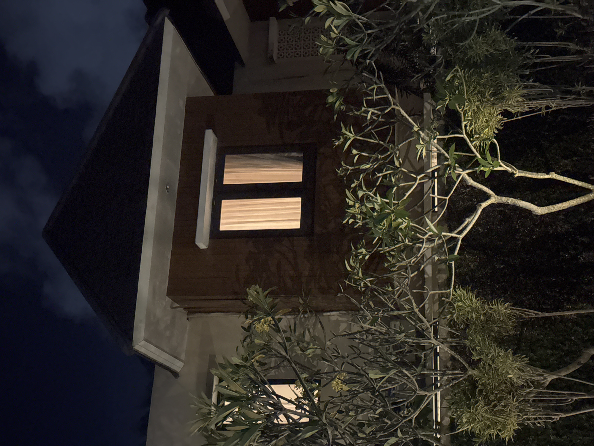 The image depicts the exterior of a multi-story building at night in Kuta Selatan, Indonesia. A section of the building's facade is visible, featuring brown wooden paneling and two rectangular windows with horizontal blinds. Both windows emit light. A dark roofline is on the left side of the frame. To the right, branches of a tree with light green leaves obscure parts of the building. The background is dark, indicating nighttime.