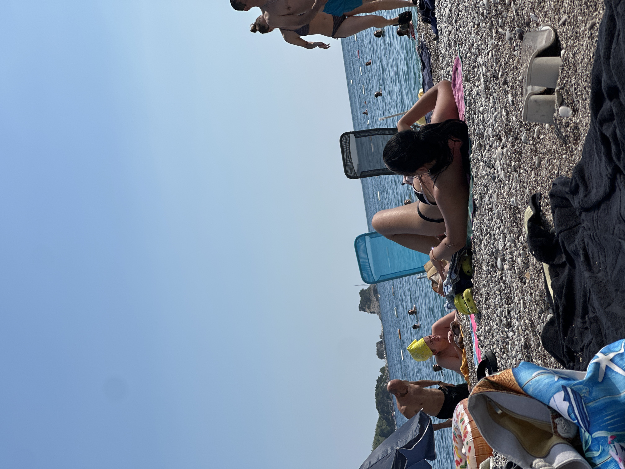 The image depicts a sunny day at the beach in Budva, Montenegro. In the foreground, a woman is lying on her stomach on a pink towel, facing away from the camera, wearing a black bikini. Her legs are crossed at the ankles. Beside her on the pebbled beach are various items including a black towel, a pair of white sandals, and what appears to be a beach bag. Further back, a man in black shorts is standing in shallow water, looking down. A woman wearing a yellow headscarf is also in the water, resting on a towel. In the background, other people are visible in the sea, and rocky islands are in the distance against a clear blue sky. Blue beach chairs are also scattered on the shore.