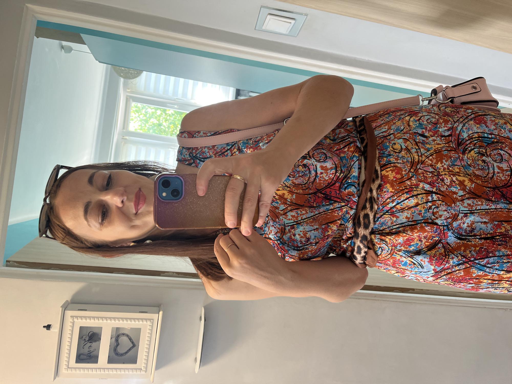 This is a portrait-oriented, eye-level, selfie taken in a mirror. A woman with long, dark brown hair and light-colored eyes is visible. She is wearing sunglasses on her head, a gold ring on her left hand, and a colorful, abstract print dress with a leopard print belt. A light pink purse is slung over her shoulder. The background shows a brightly lit room with a window, suggesting it is daytime. There is a white framed object on the wall below the mirror, which appears to be a decorative box or jewelry organizer with two compartments, each containing a heart-shaped item. The overall mood is casual and relaxed.
