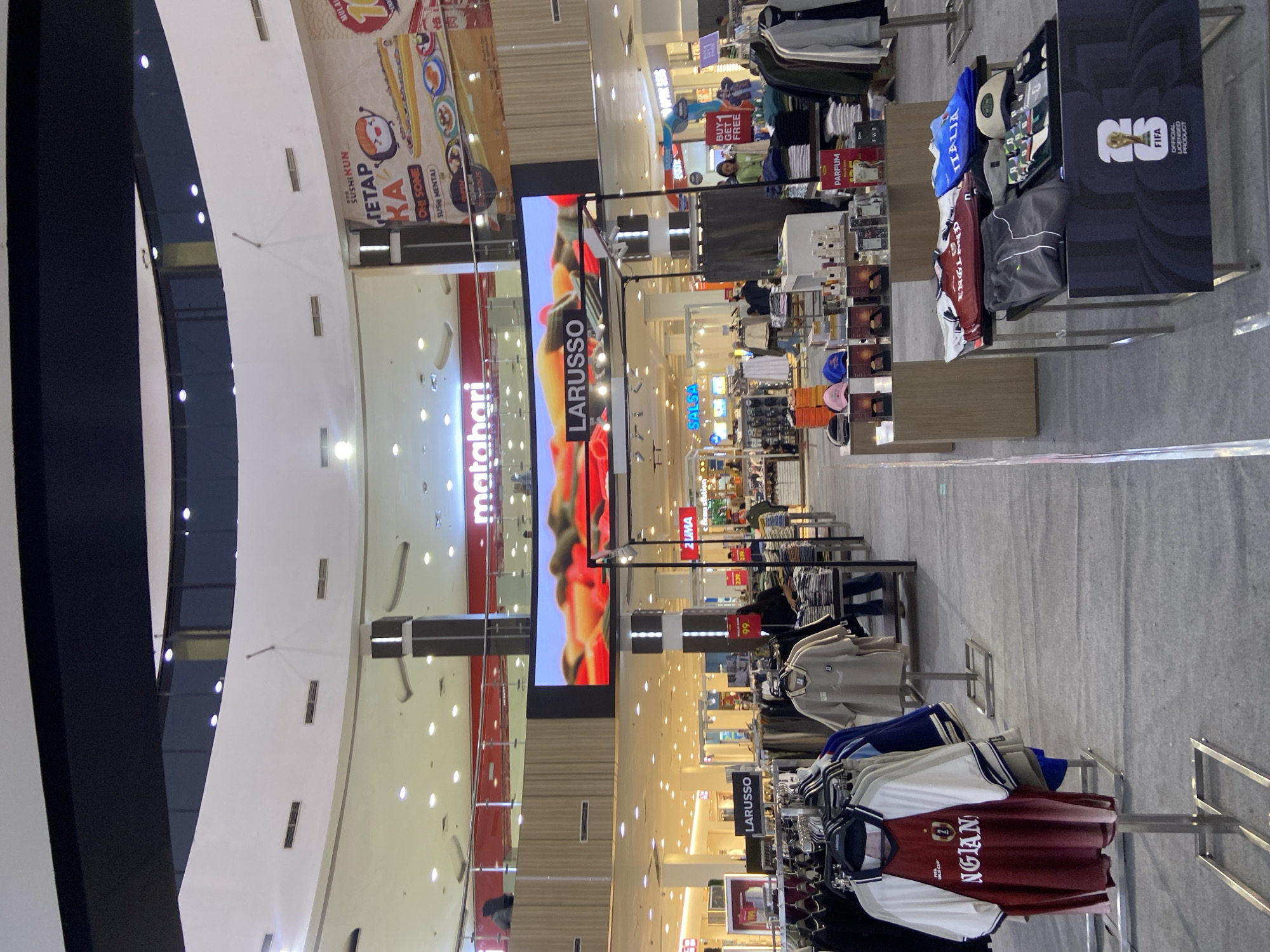 The image depicts the interior of a shopping mall, likely the Sun City Mall in Sidoarjo, Indonesia. A prominent digital billboard displays the "LARUSSO" brand name above a vibrant image of pasta. To the left, a "Matahari" department store sign is visible. Racks of clothing are displayed for sale, including t-shirts, jackets, and sportswear. A person is seen browsing a clothing rack, and another is standing near a display of perfumes. The mall features a multi-level design with escalators and walkways. The overall lighting is bright and commercial, indicative of a retail environment.