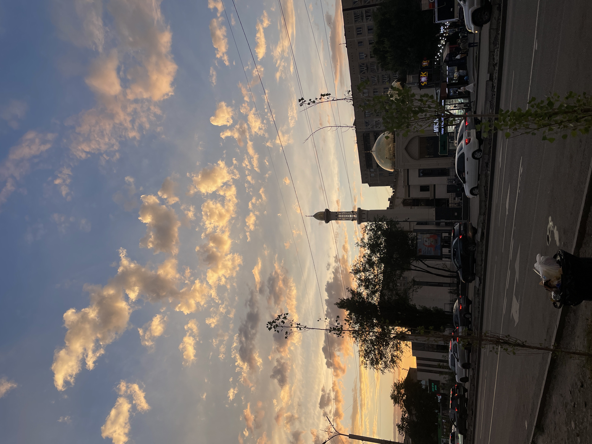 The image depicts a street scene in Almaty, Kazakhstan, during sunset. The sky is filled with dramatic clouds tinged with orange and yellow hues from the setting sun, contrasting with the clear blue sky above. In the middle ground, a mosque with a golden dome and a minaret is visible, flanked by trees and buildings. A multi-lane road runs across the foreground, with several cars driving on it, some with their headlights on. A trash can overflowing with refuse is positioned on the sidewalk on the right side of the frame, near a young tree. Overhead power lines are also visible, strung across the sky. The overall impression is an urban landscape at dusk, with a prominent religious structure and the everyday activity of city life.