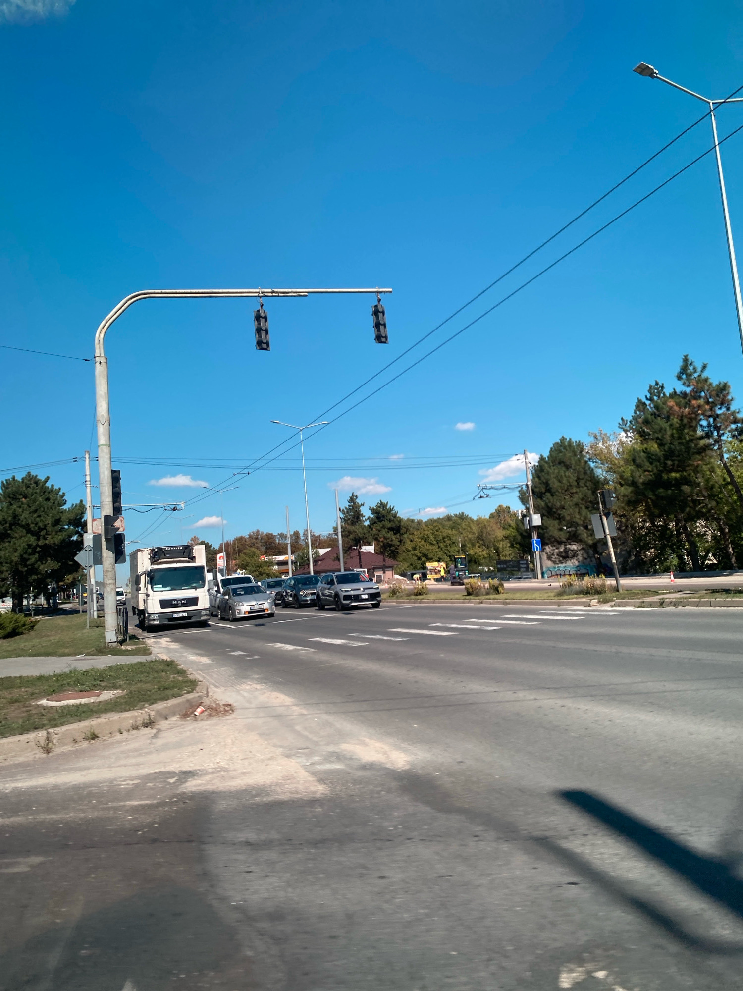 The image captures a sunny day at a traffic intersection in Chișinău, Moldova. The sky is a clear, bright blue with a few wispy white clouds. A large traffic light hangs over the intersection from a metal pole. In the foreground, the road shows white pedestrian crossings. Several vehicles are present, including a white truck with the "MAN" logo and "CARRIER" branding on its cooler unit, several cars, and in the distance, a yellow construction vehicle. Trees line the sides of the road, and houses are visible in the background. The scene appears to be a typical urban streetscape.
