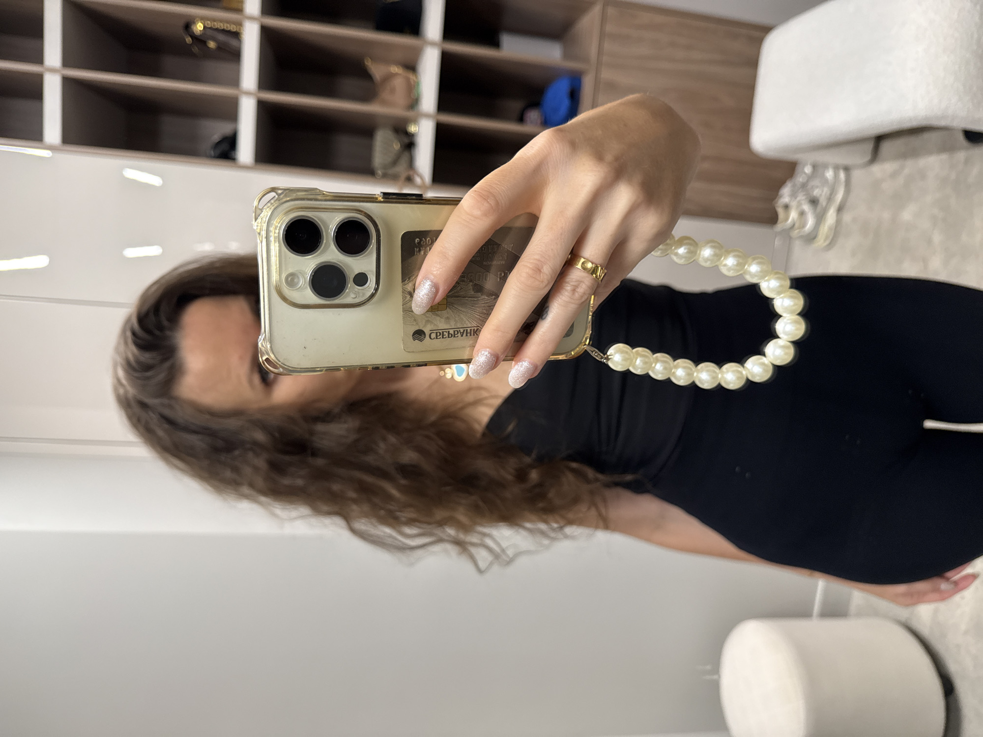 In Moscow, Russia, a woman takes a mirror selfie in what appears to be a dressing room or closet. She is wearing a black top and black pants and is holding a smartphone with a pearl charm attached. In the background, shelves are visible, containing various items, including handbags and shoes. A white ottoman is also present in the room.