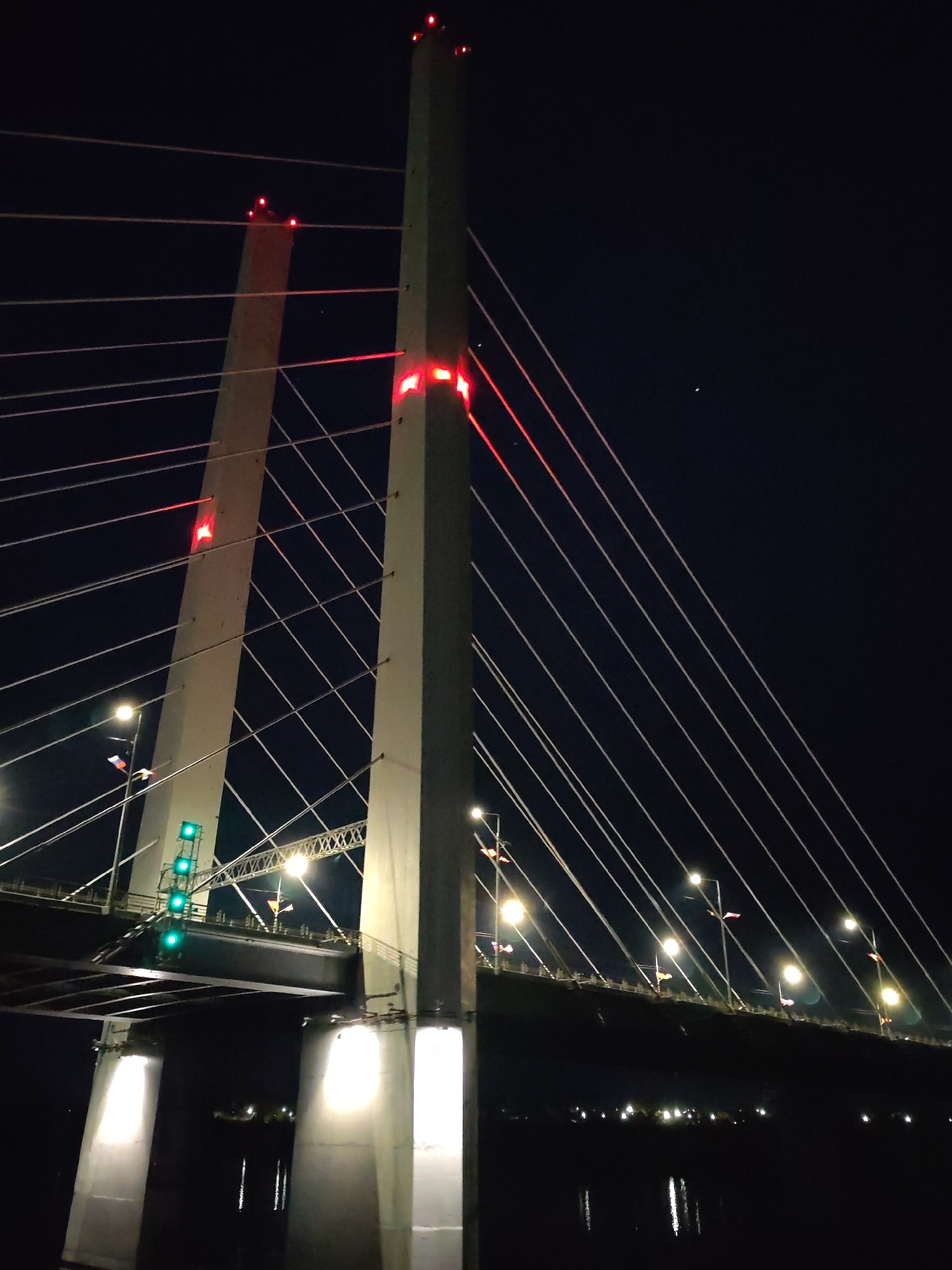 The image depicts the October Bridge in Cherepovets, Russia, at night. The bridge is a cable-stayed structure with two prominent pylons. Both pylons are illuminated with red lights at their tops and mid-sections. The deck of the bridge is visible, with streetlights along its length. Support pillars are brightly lit at the base. Numerous cables stretch from the pylons to the bridge deck. Some traffic lights are present on the bridge, shining green. The background is a dark night sky, with a faint celestial object visible in the upper right. The water below reflects some of the bridge's lights.