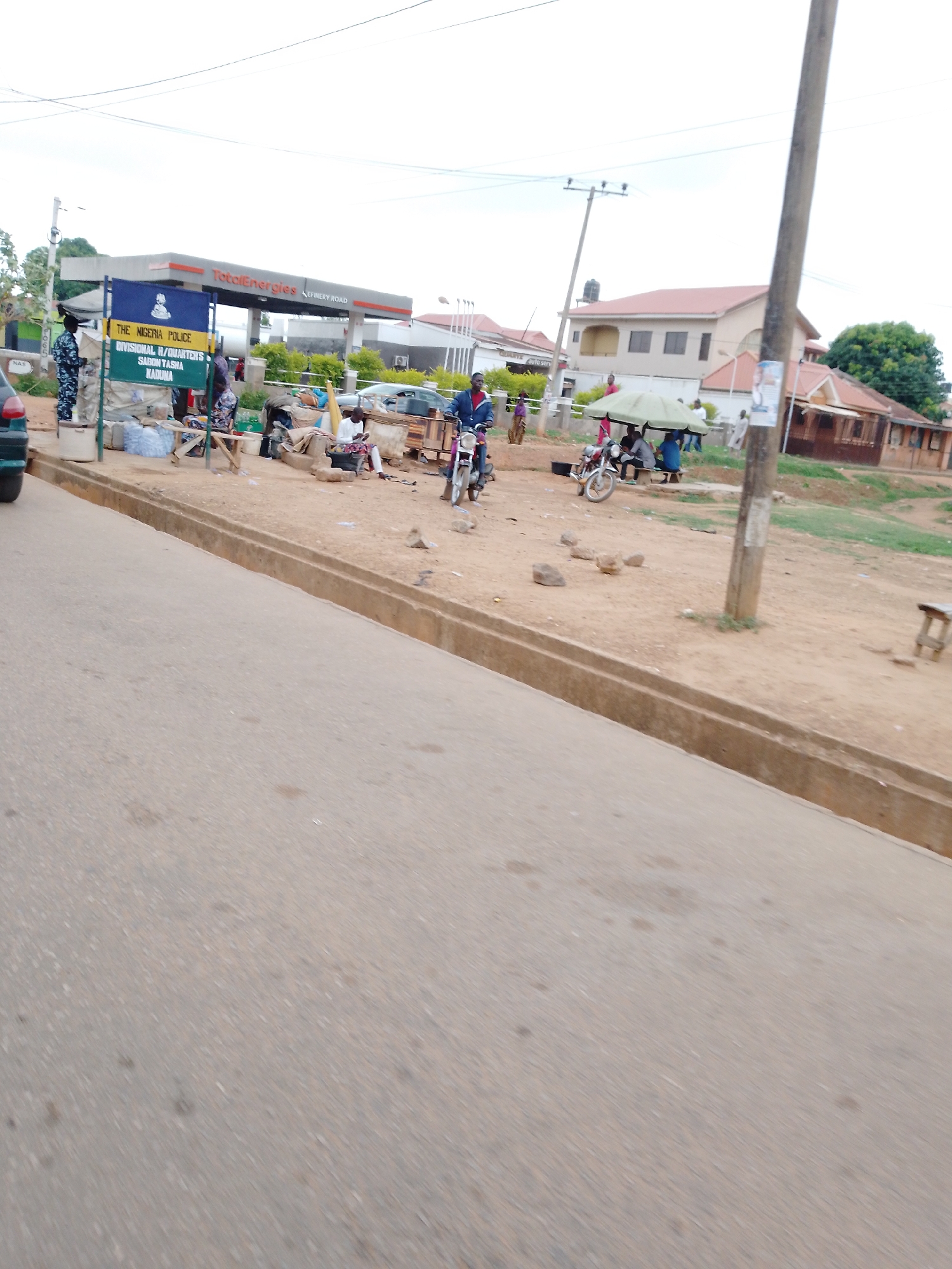 The image depicts a street scene in Sabon Tasha, Kaduna, Nigeria. A TotalEnergies gas station is visible in the background, alongside a sign for "The Nigeria Police Divisional H/Quarters Sabon Tasha Kaduna." A police officer in uniform stands near the sign. Several individuals are present along the roadside, some seated and others on motorcycles. The road appears to be paved. There are also poles in the image.